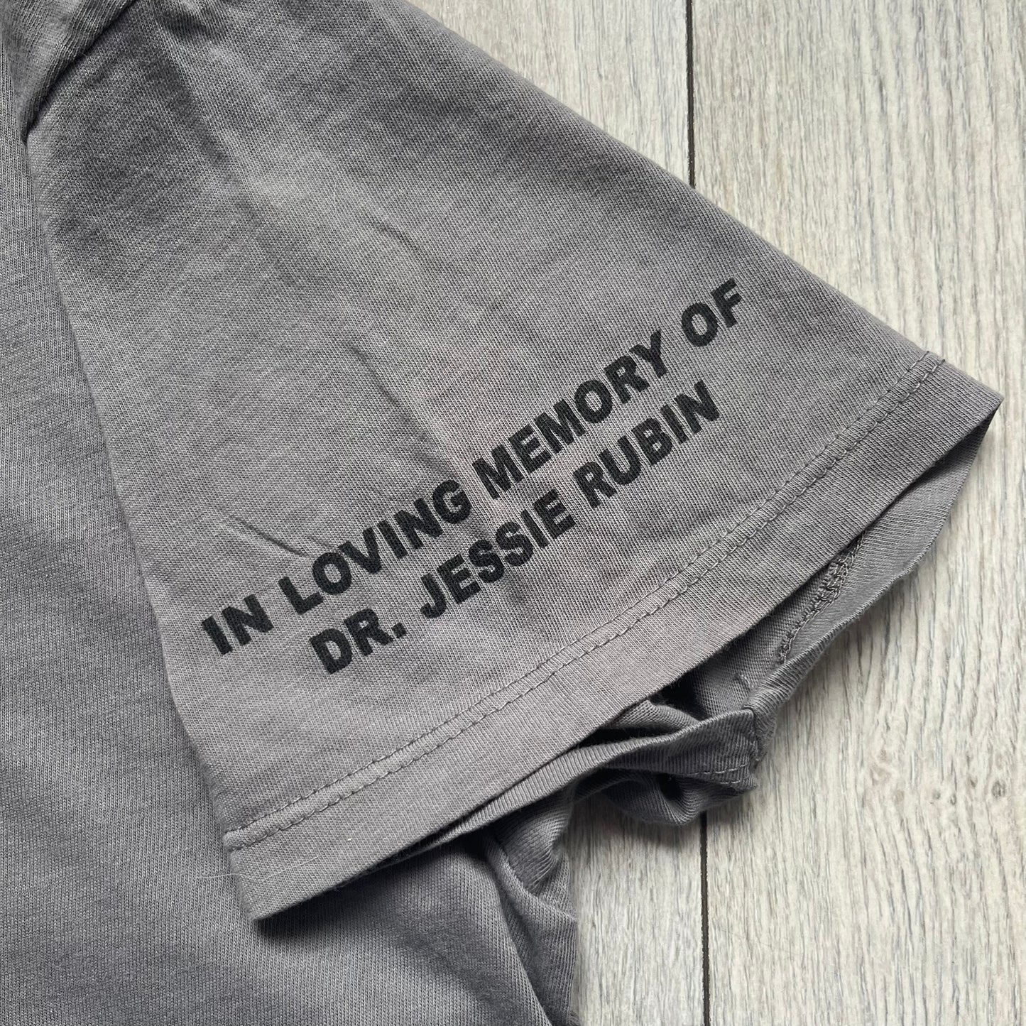 Marc Jacobs 2010s Robert Duffy Skin Cancer Awareness T-shirt is a gray campaign tee created to support skin cancer awareness. The front features an illustrative composition of Robert Duffy with Marc Jacobs included in the design. The sleeves contain printed text referencing “In loving memory of Dr Jessie Rubin” and support for NYU School of Medicine. The back includes a large “Thank You” message listing contributors involved in the campaign.
