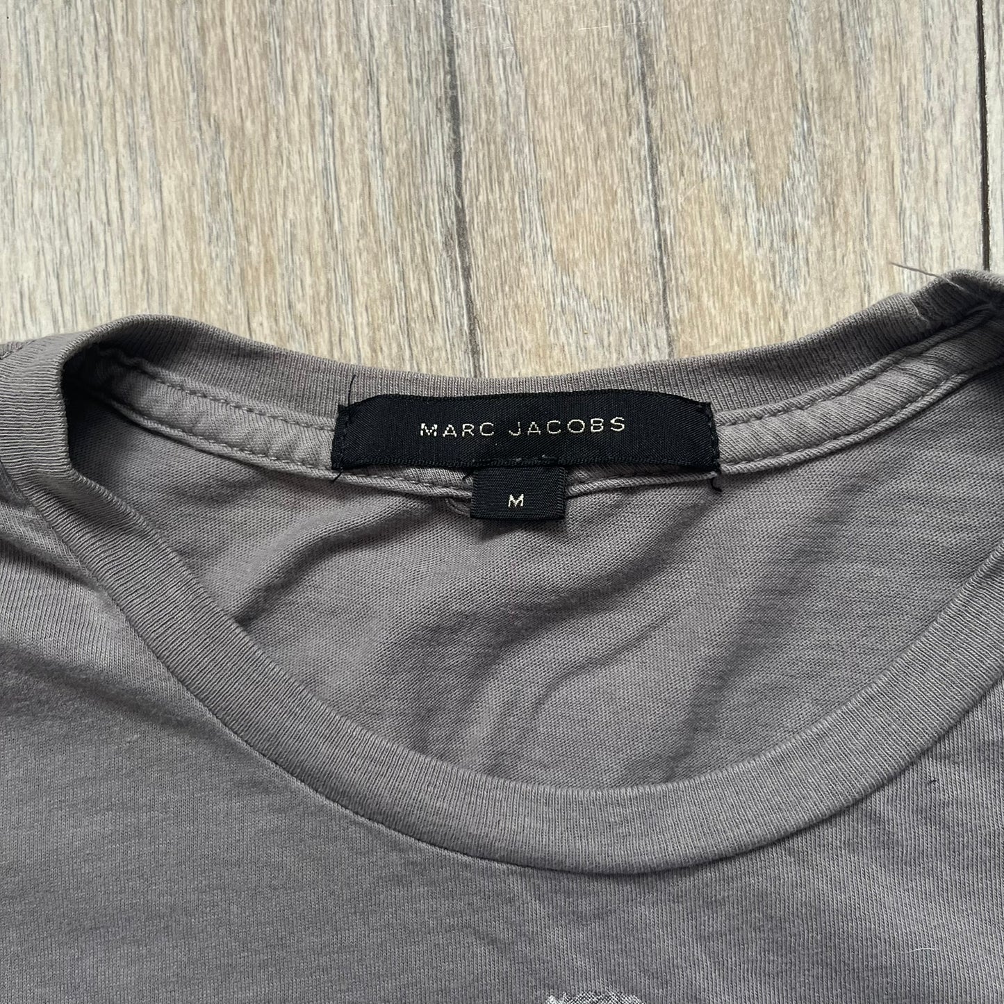 Marc Jacobs 2010s Robert Duffy Skin Cancer Awareness T-shirt is a gray campaign tee created to support skin cancer awareness. The front features an illustrative composition of Robert Duffy with Marc Jacobs included in the design. The sleeves contain printed text referencing “In loving memory of Dr Jessie Rubin” and support for NYU School of Medicine. The back includes a large “Thank You” message listing contributors involved in the campaign.
