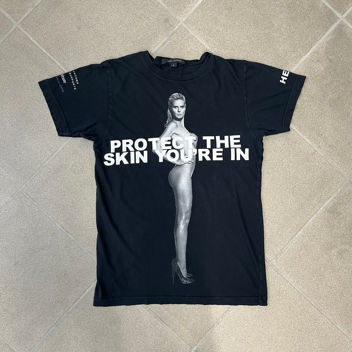Marc Jacobs 2010s “Protect The Skin You’re In” Heidi Klum T-shirt is a black campaign tee created for skin cancer awareness. The front features an illustrative image of Heidi Klum with “Protect The Skin You’re In” text integrated into the design. The sleeves include "Heidi” and support for NYU Medical Center Cancer Institute. The back displays a large “Thank You” message listing contributors to the campaign.