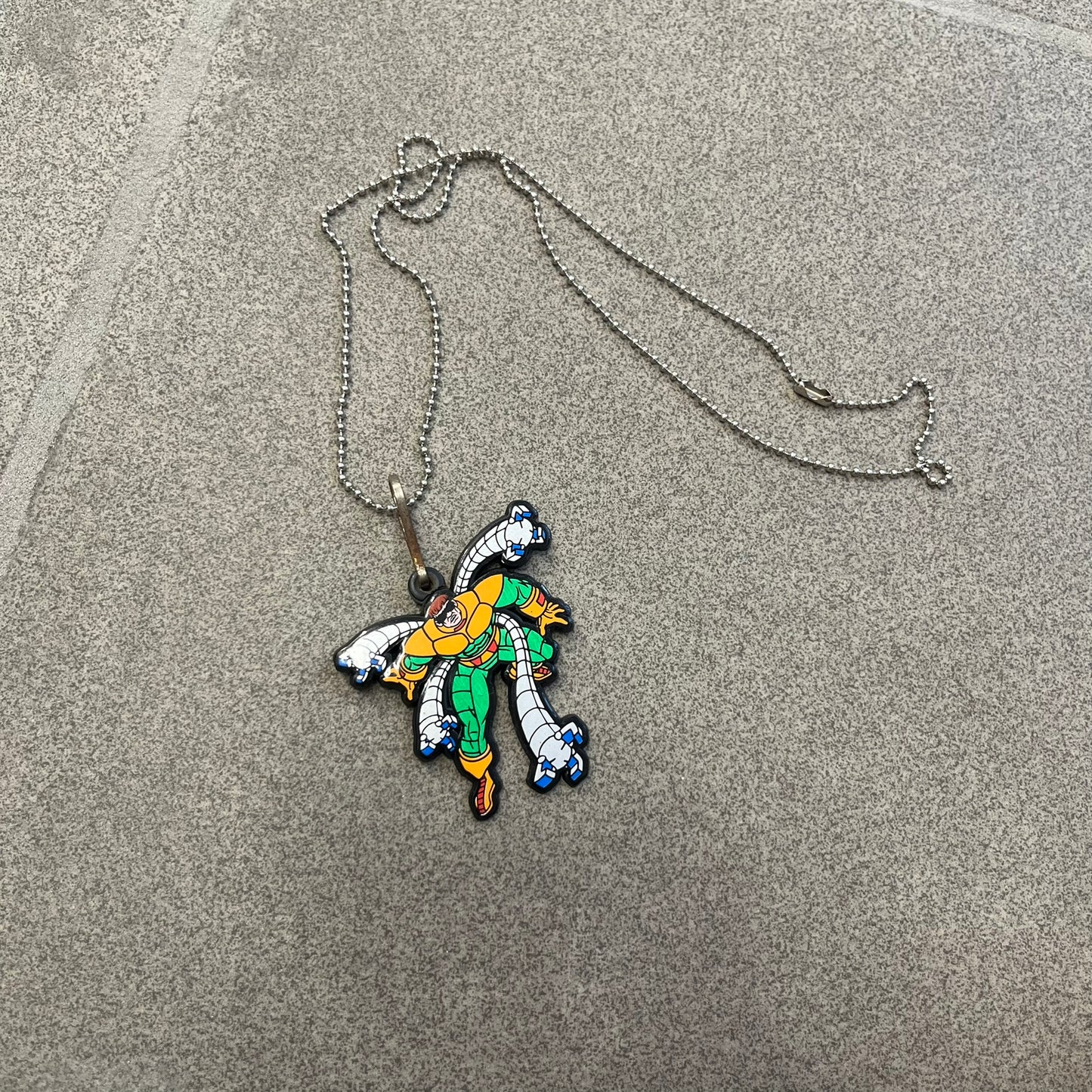 Marvel 1995 Doctor Octopus Necklace features a 2D cartoon-style Doctor Octopus pendant with a colorful graphic on the front. The back of the pendant is black and includes 1995 Marvel licensing details. It has a metal loop at the top and comes with a basic steel chain.
