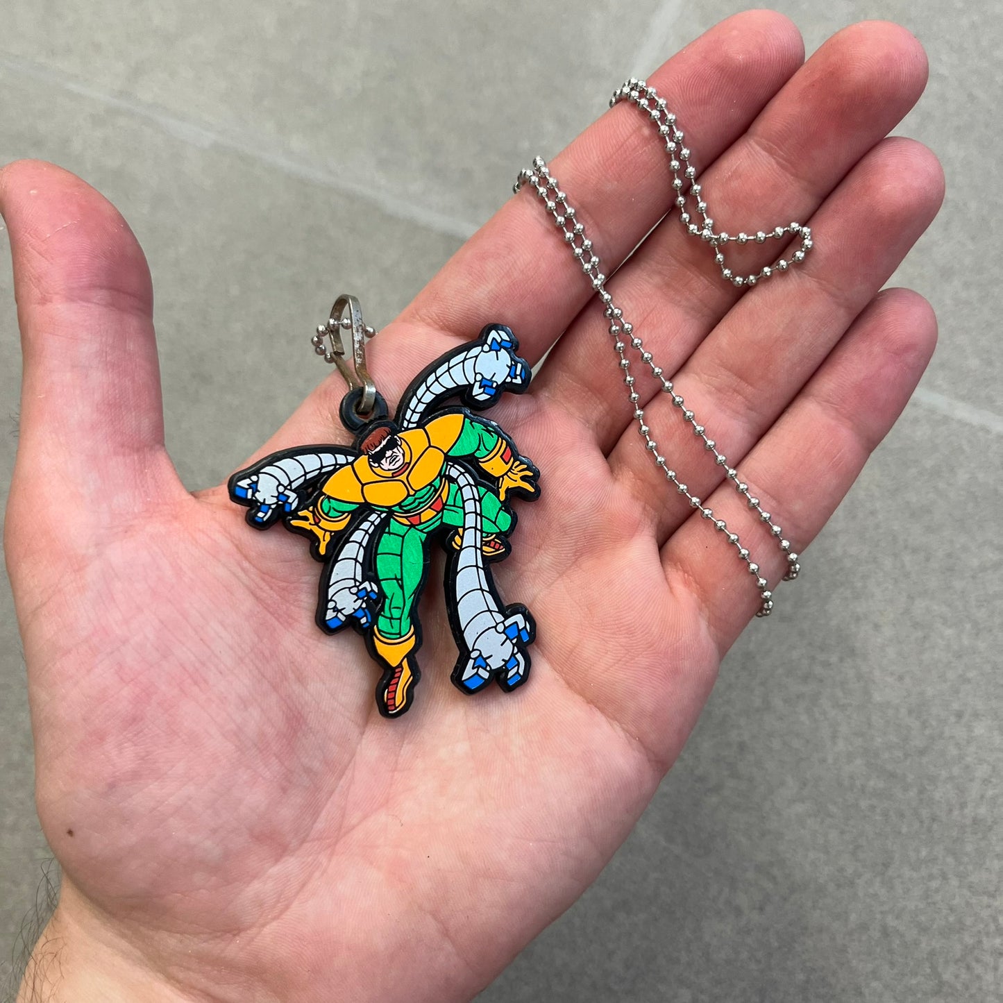 Marvel 1995 Doctor Octopus Necklace features a 2D cartoon-style Doctor Octopus pendant with a colorful graphic on the front. The back of the pendant is black and includes 1995 Marvel licensing details. It has a metal loop at the top and comes with a basic steel chain.

