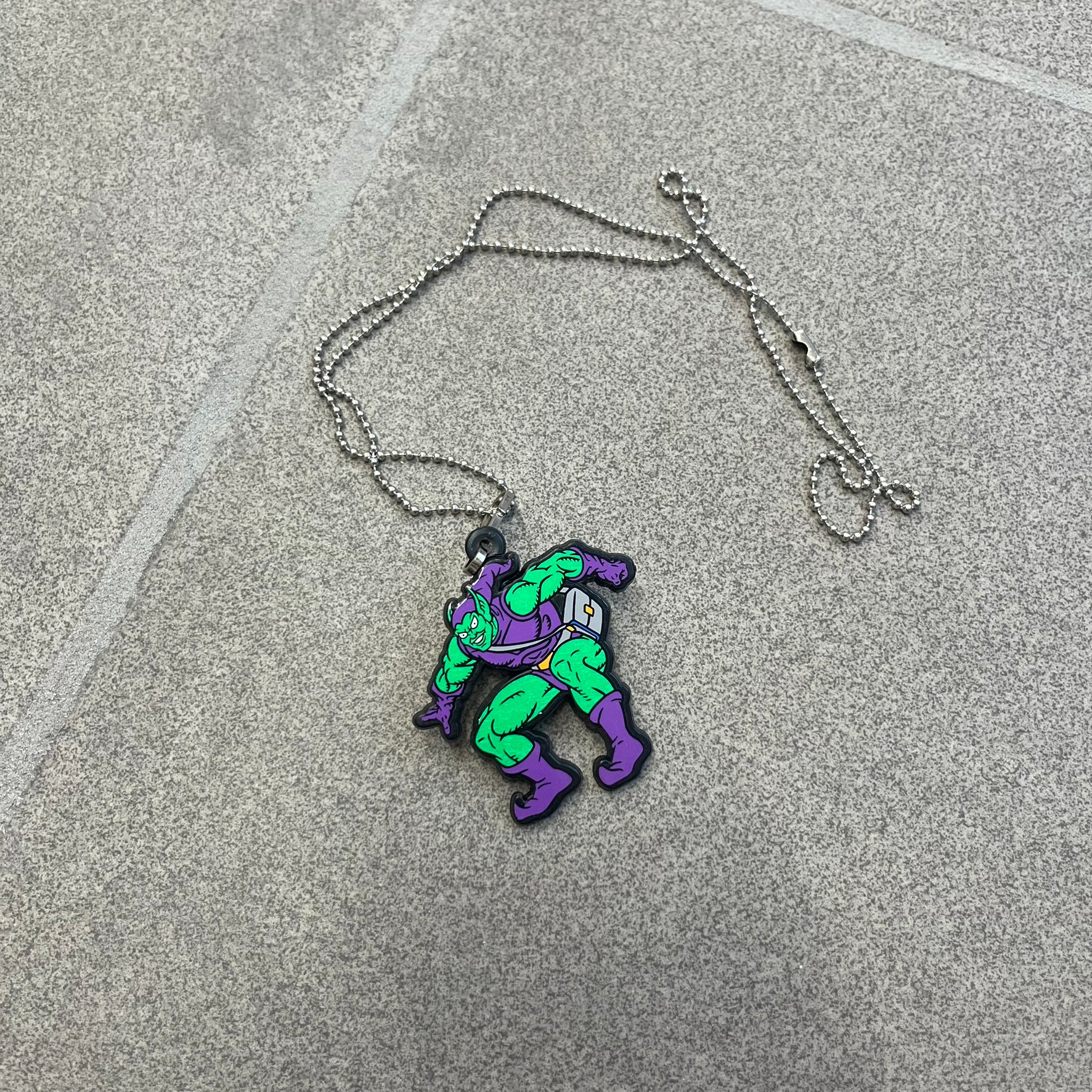 Marvel 1995 Green Goblin Necklace features a 2D cartoon-style Green Goblin pendant with a colorful graphic on the front. The back of the pendant is black and includes 1995 Marvel licensing details. It has a metal loop at the top and comes with a basic steel chain.