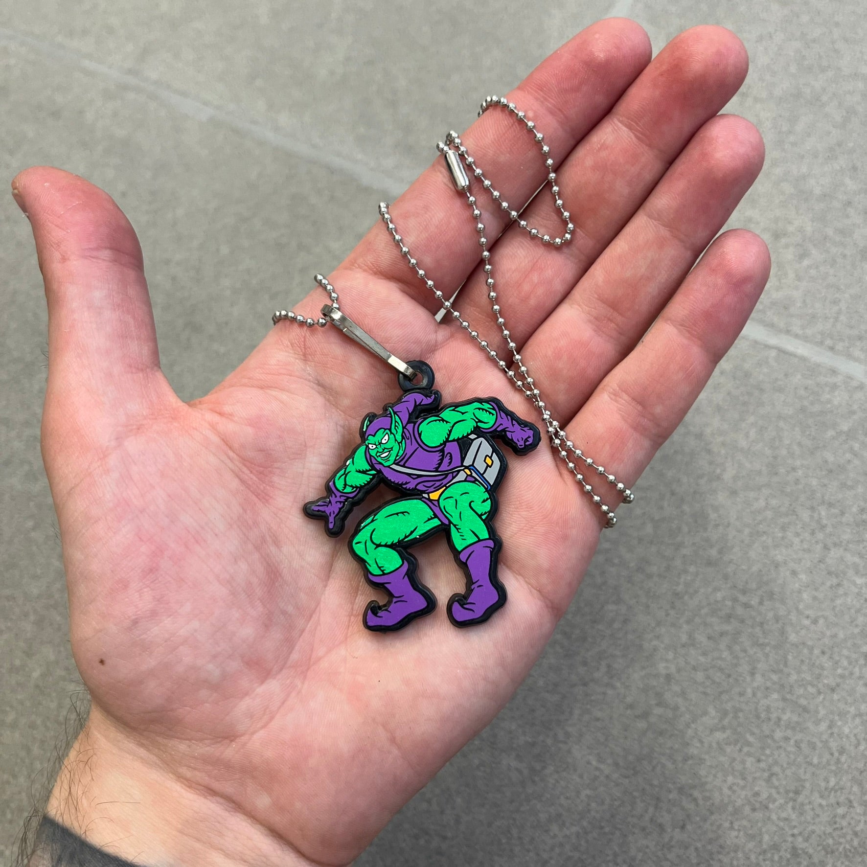 Marvel 1995 Green Goblin Necklace features a 2D cartoon-style Green Goblin pendant with a colorful graphic on the front. The back of the pendant is black and includes 1995 Marvel licensing details. It has a metal loop at the top and comes with a basic steel chain.