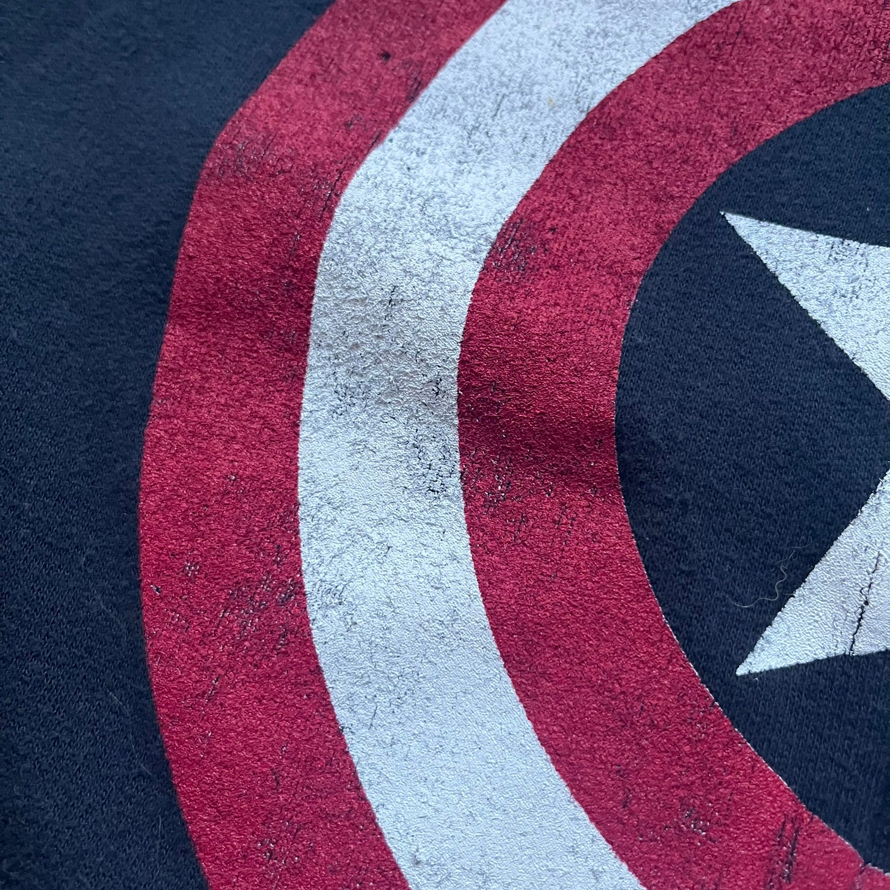 Black hoodie with a large Captain America shield graphic on the front. Made from an 80/20 polyester blend, lightweight. Features metal zipper, front pockets, and WeLoveFine × Marvel neck tag. The shield graphic has an intentional rough texture.