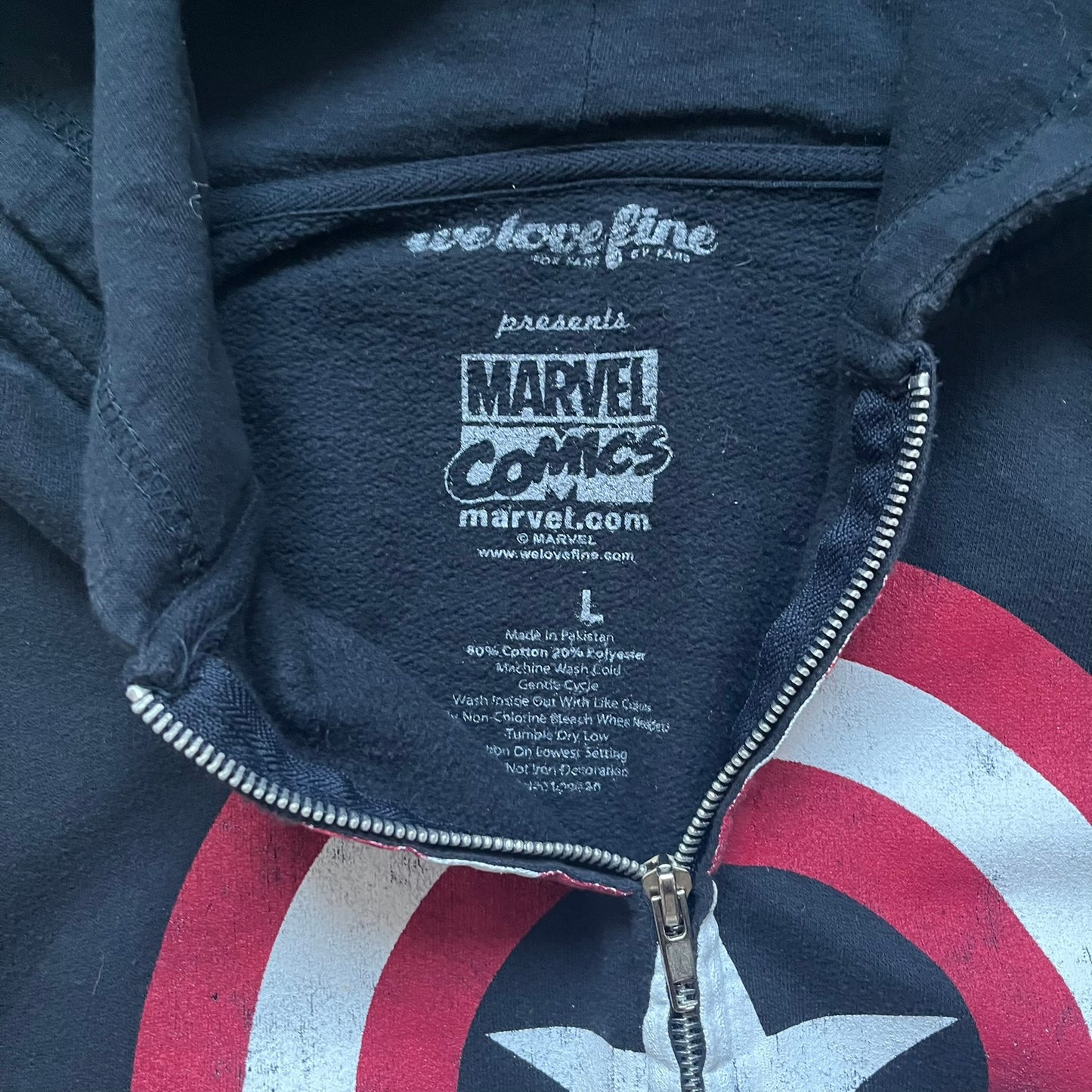 Black hoodie with a large Captain America shield graphic on the front. Made from an 80/20 polyester blend, lightweight. Features metal zipper, front pockets, and WeLoveFine × Marvel neck tag. The shield graphic has an intentional rough texture.