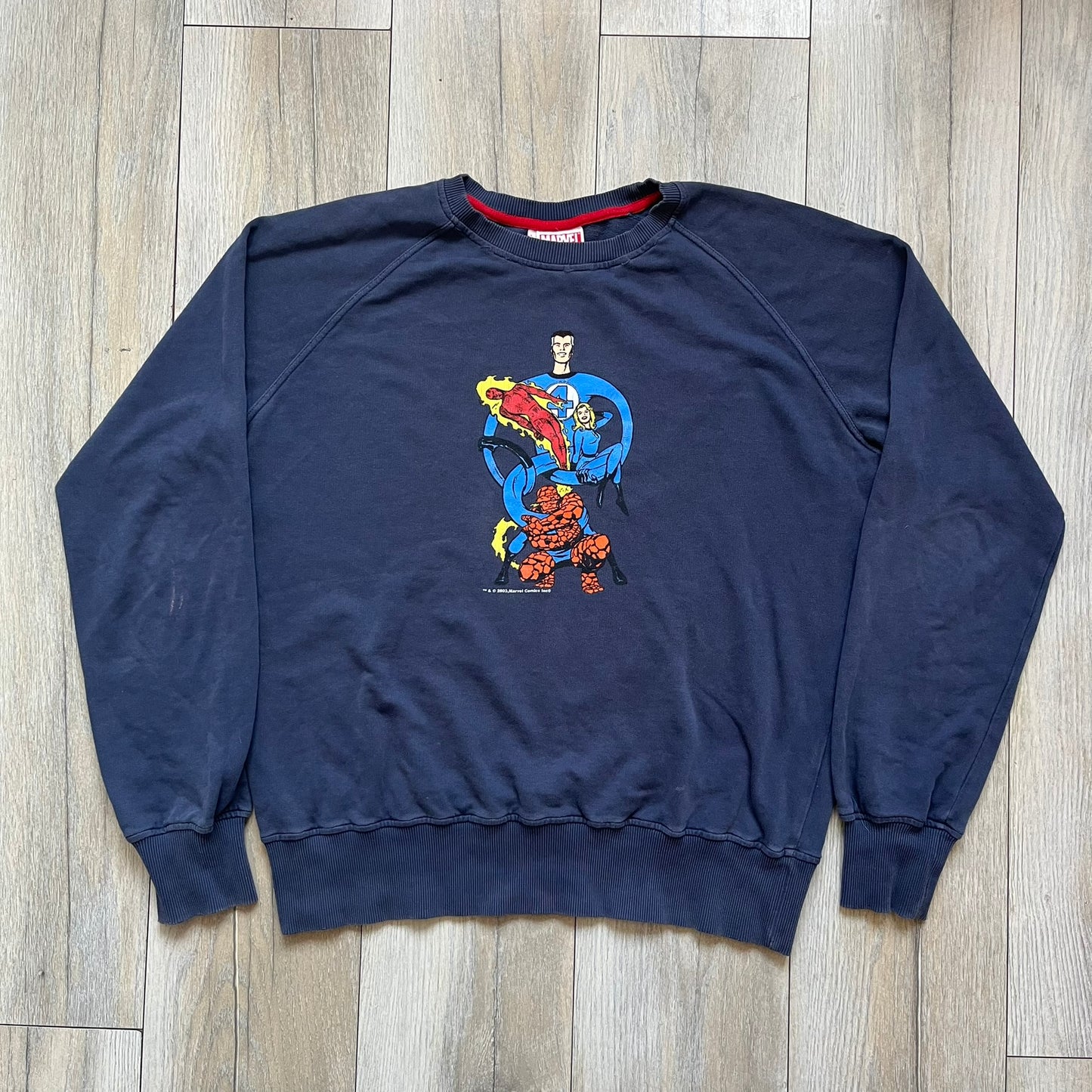 Marvel Fantastic Four 2003 Navy Crewneck is a navy sweatshirt made from 100% cotton with an official Marvel neck tag. The front features a central graphic of the Fantastic Four characters intertwined together, with Reed Richards in a stretched form, Human Torch flying above, Ben Grimm holding Reed, and Sue Storm positioned within the composition. Small white licensing text with the 2003 date appears beneath the graphic.