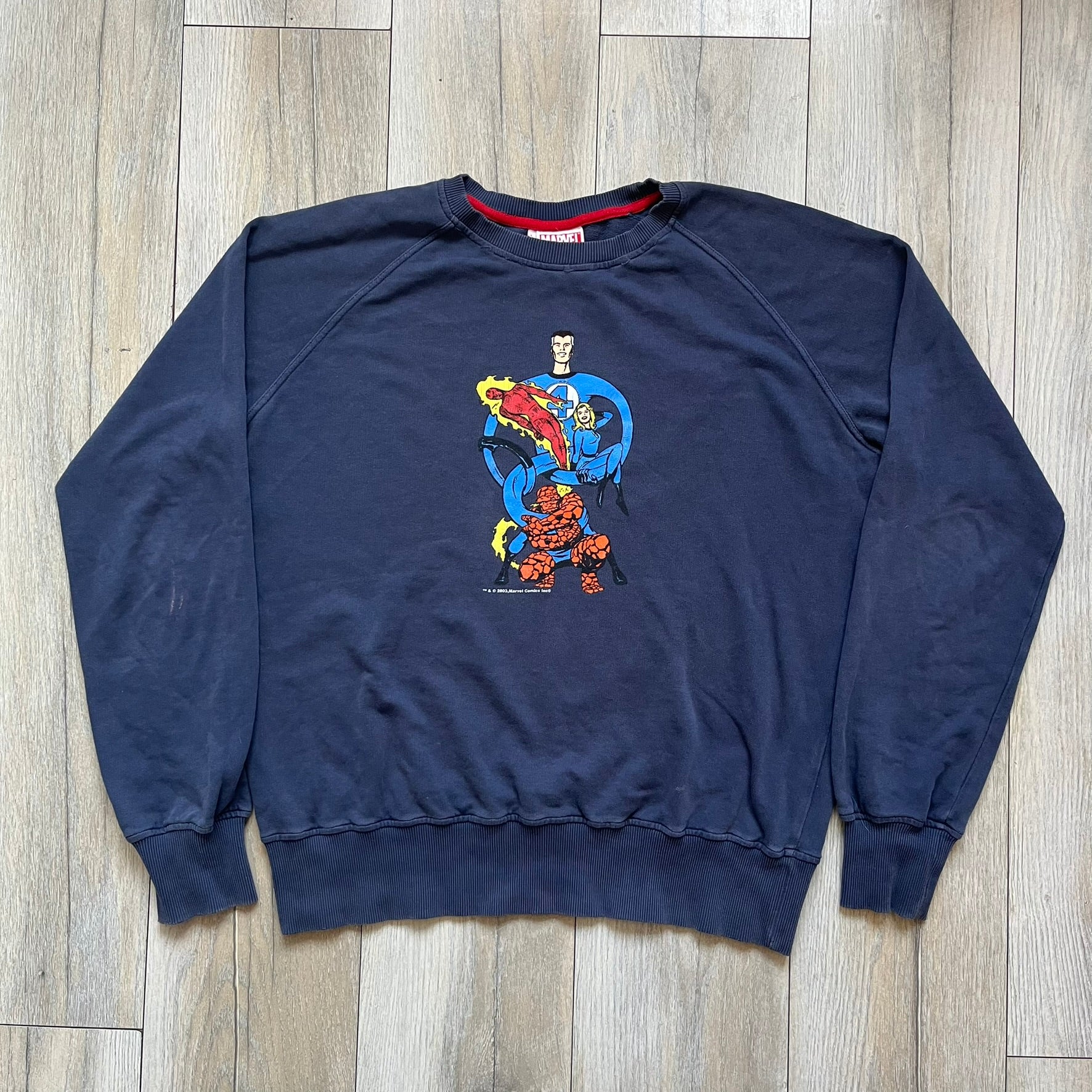 Marvel Fantastic Four 2003 Navy Crewneck is a navy sweatshirt made from 100% cotton with an official Marvel neck tag. The front features a central graphic of the Fantastic Four characters intertwined together, with Reed Richards in a stretched form, Human Torch flying above, Ben Grimm holding Reed, and Sue Storm positioned within the composition. Small white licensing text with the 2003 date appears beneath the graphic.