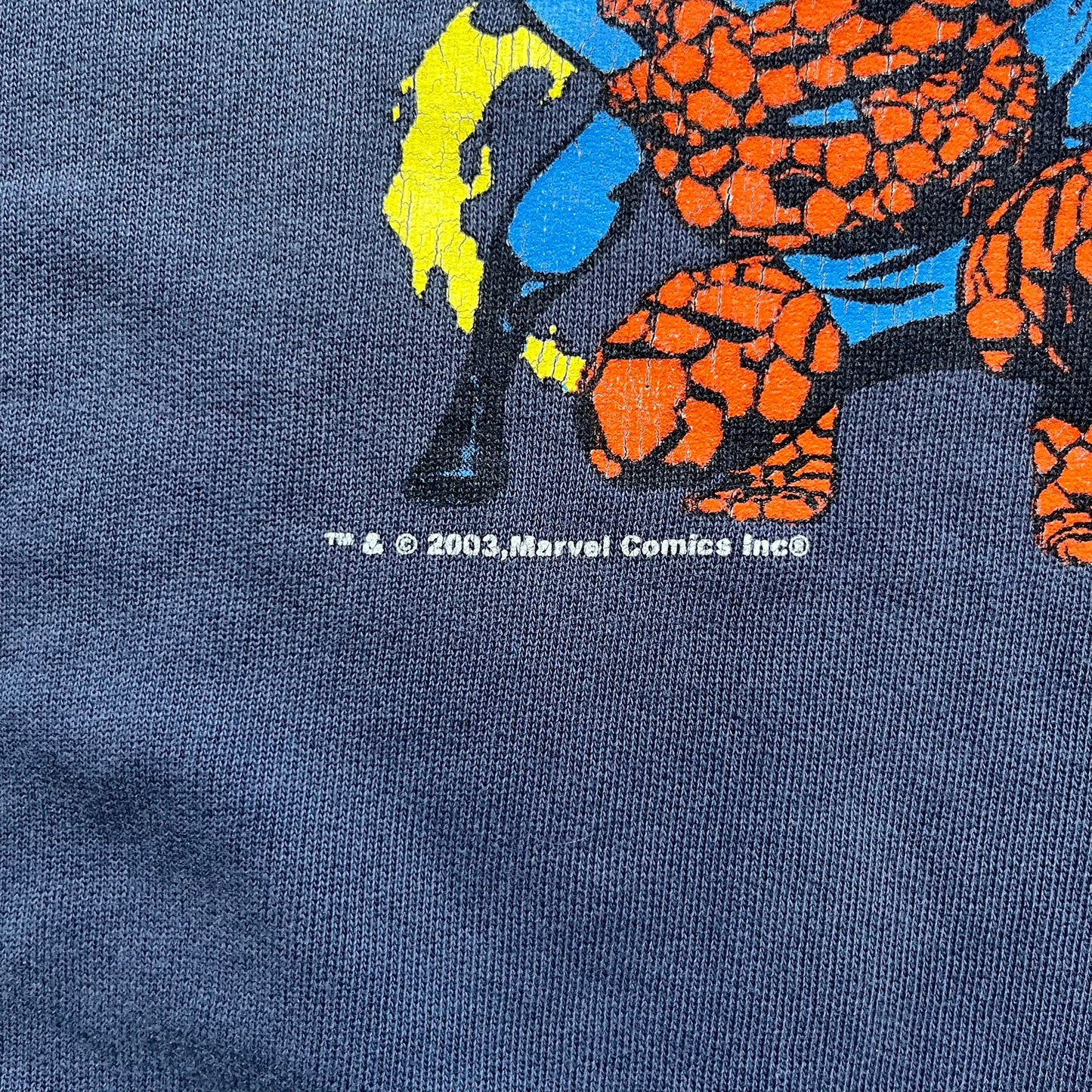 Marvel Fantastic Four 2003 Navy Crewneck is a navy sweatshirt made from 100% cotton with an official Marvel neck tag. The front features a central graphic of the Fantastic Four characters intertwined together, with Reed Richards in a stretched form, Human Torch flying above, Ben Grimm holding Reed, and Sue Storm positioned within the composition. Small white licensing text with the 2003 date appears beneath the graphic.