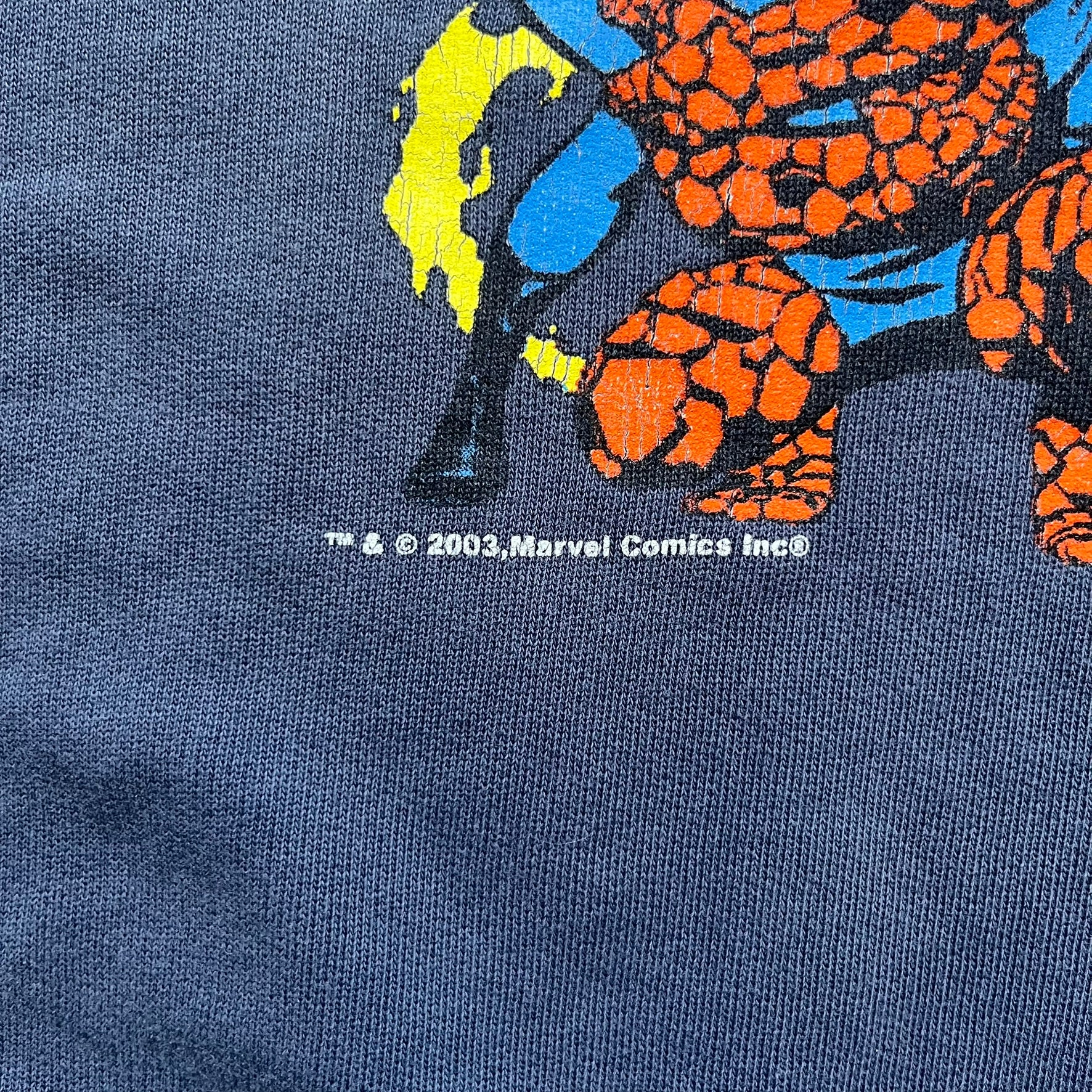 Marvel Fantastic Four 2003 Navy Crewneck is a navy sweatshirt made from 100% cotton with an official Marvel neck tag. The front features a central graphic of the Fantastic Four characters intertwined together, with Reed Richards in a stretched form, Human Torch flying above, Ben Grimm holding Reed, and Sue Storm positioned within the composition. Small white licensing text with the 2003 date appears beneath the graphic.