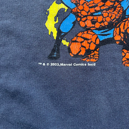 Marvel Fantastic Four 2003 Navy Crewneck is a navy sweatshirt made from 100% cotton with an official Marvel neck tag. The front features a central graphic of the Fantastic Four characters intertwined together, with Reed Richards in a stretched form, Human Torch flying above, Ben Grimm holding Reed, and Sue Storm positioned within the composition. Small white licensing text with the 2003 date appears beneath the graphic.