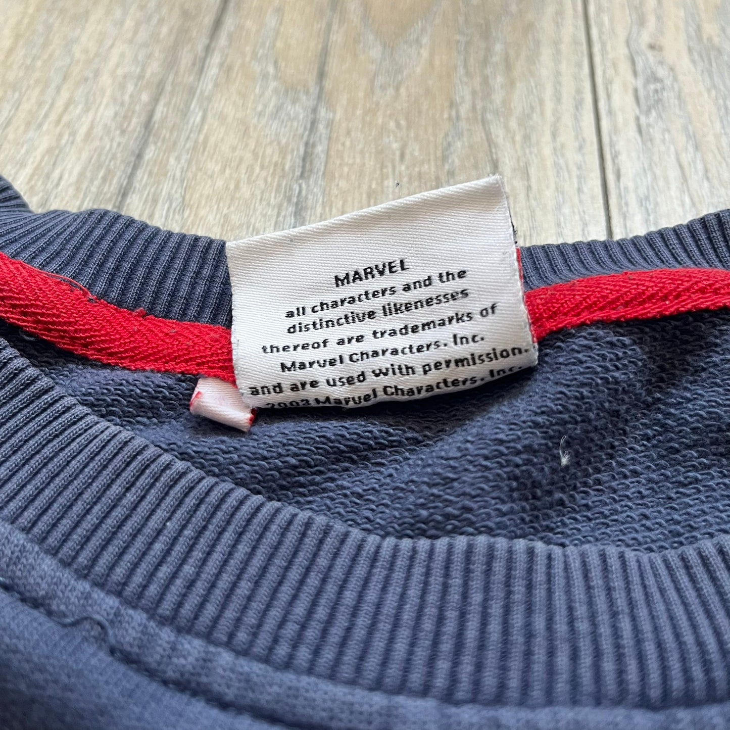 Marvel Fantastic Four 2003 Navy Crewneck is a navy sweatshirt made from 100% cotton with an official Marvel neck tag. The front features a central graphic of the Fantastic Four characters intertwined together, with Reed Richards in a stretched form, Human Torch flying above, Ben Grimm holding Reed, and Sue Storm positioned within the composition. Small white licensing text with the 2003 date appears beneath the graphic.