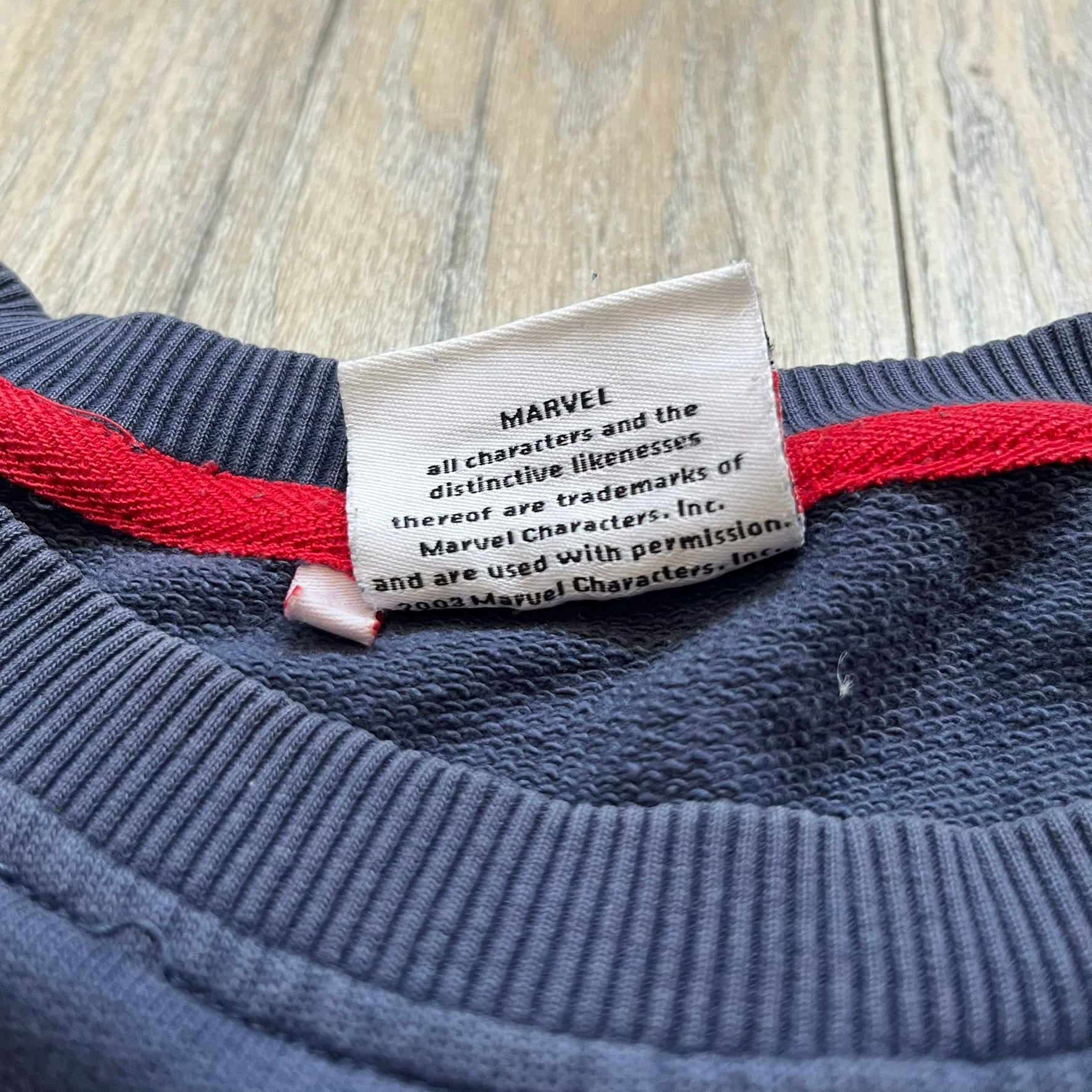 Marvel Fantastic Four 2003 Navy Crewneck is a navy sweatshirt made from 100% cotton with an official Marvel neck tag. The front features a central graphic of the Fantastic Four characters intertwined together, with Reed Richards in a stretched form, Human Torch flying above, Ben Grimm holding Reed, and Sue Storm positioned within the composition. Small white licensing text with the 2003 date appears beneath the graphic.