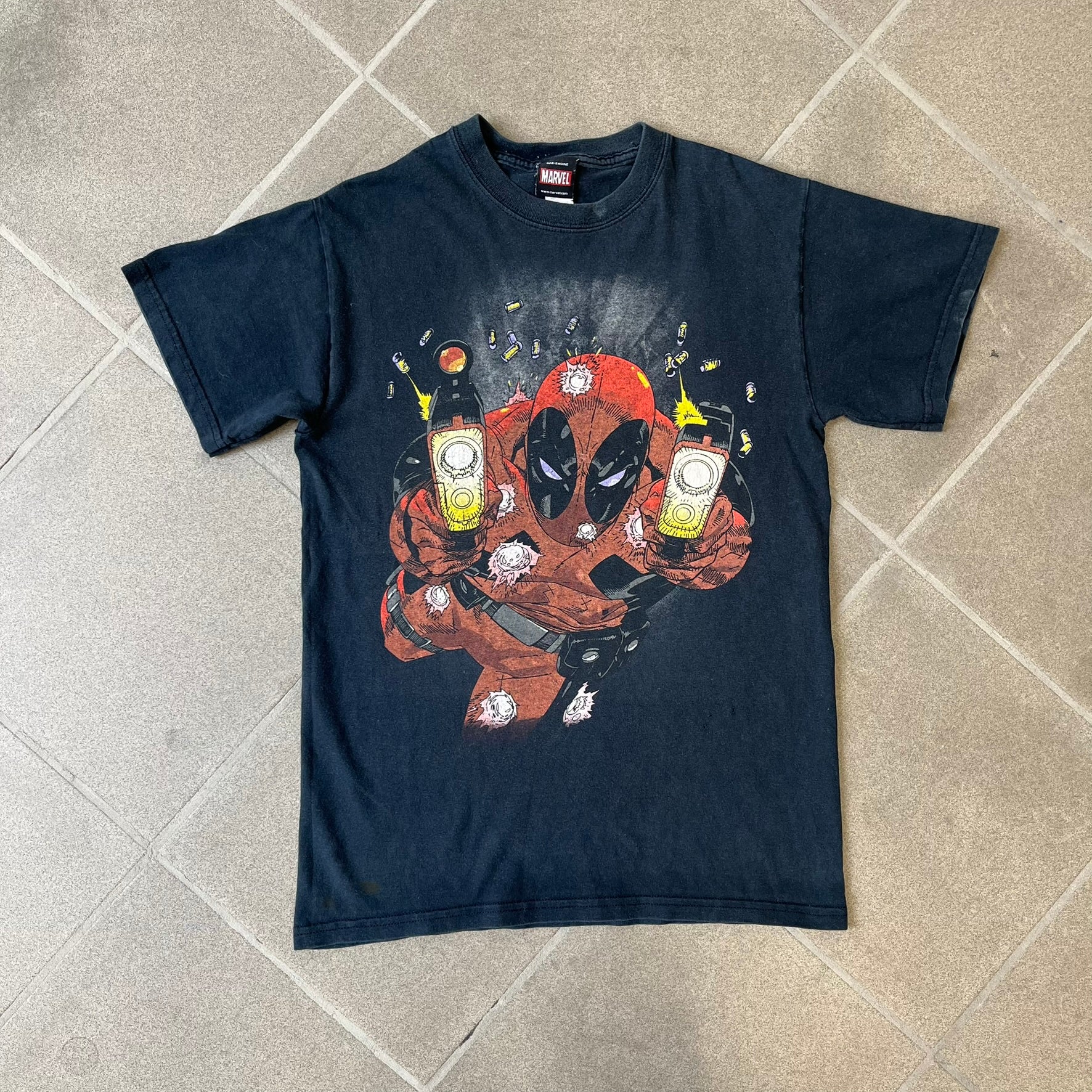 Marvel Mad Engine 2000's Deadpool Tee features a black T-shirt made by Mad Engine. The front has a comic-style graphic of Deadpool in mid-air firing two pistols, with bullets and bullet holes shown throughout the design.
