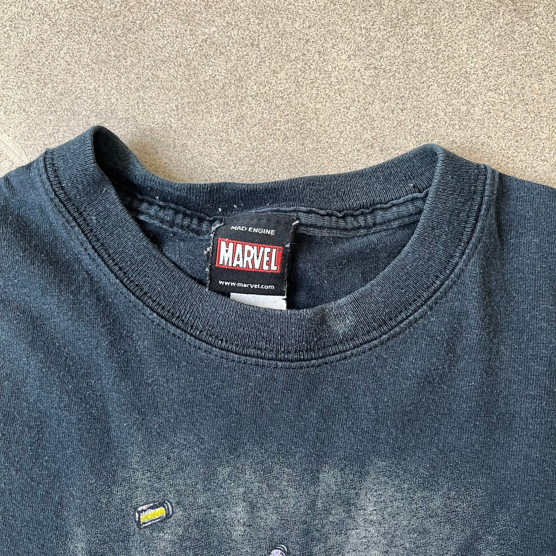Marvel Mad Engine 2000's Deadpool Tee features a black T-shirt made by Mad Engine. The front has a comic-style graphic of Deadpool in mid-air firing two pistols, with bullets and bullet holes shown throughout the design.
