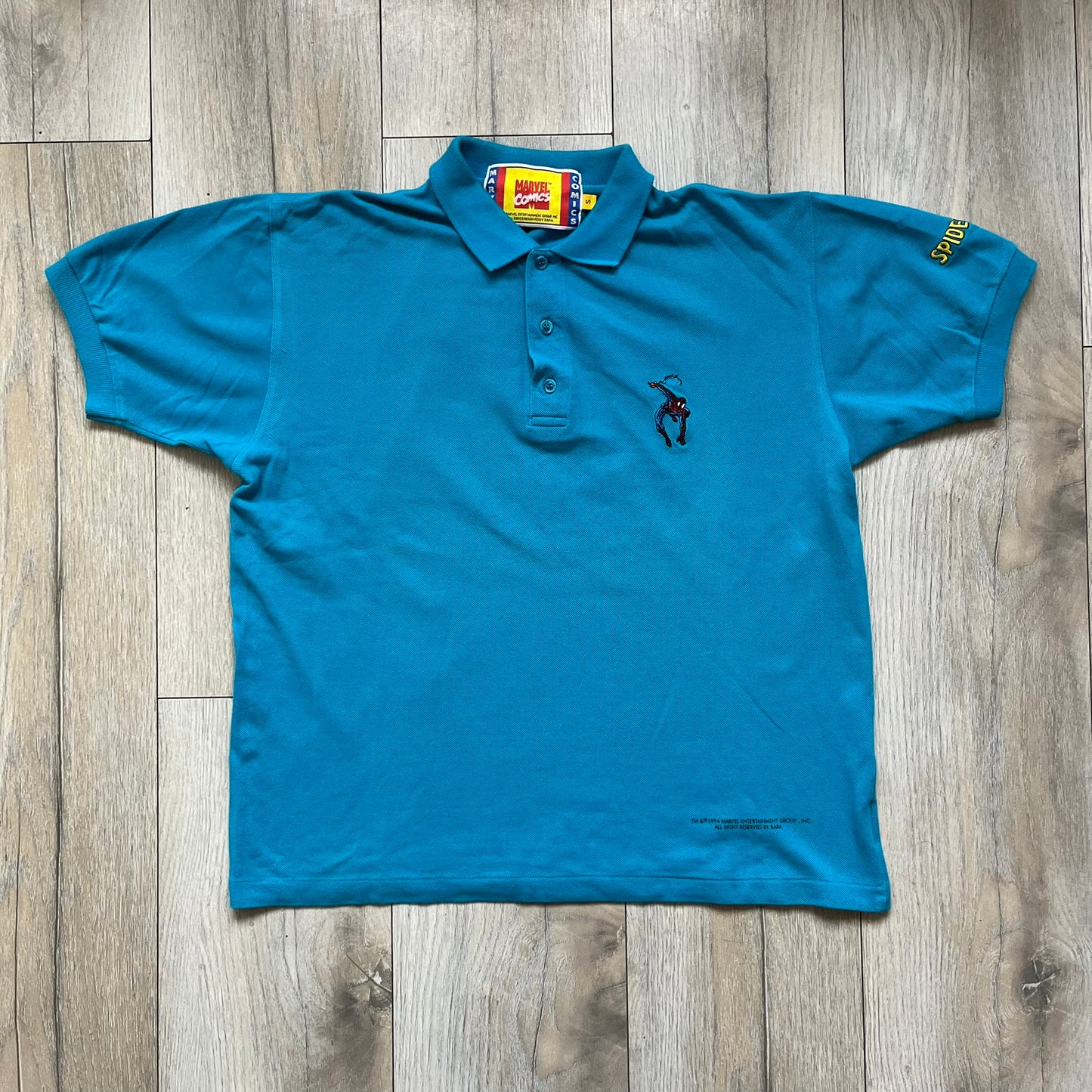 Marvel Comics Spider-Man 1994 Blue Embroidered Polo is a blue polo shirt featuring embroidered Spider-Man branding. The left chest displays an embroidered Spider-Man graphic, while the sleeve includes yellow “Spider-Man” text embroidery. The lower front is finished with small black “1994” text, and the shirt includes a large Marvel Comics neck tag.