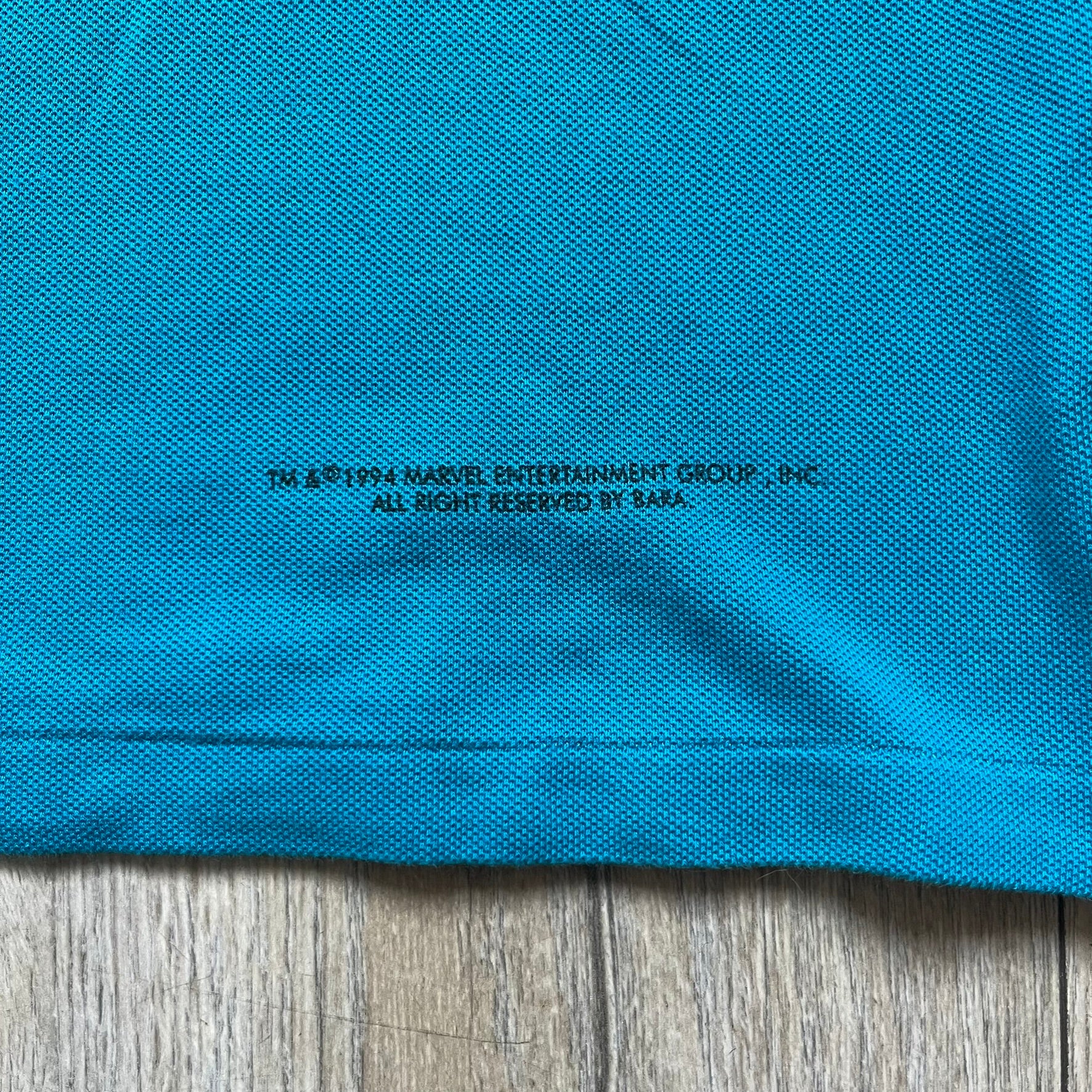 Marvel Comics Spider-Man 1994 Blue Embroidered Polo is a blue polo shirt featuring embroidered Spider-Man branding. The left chest displays an embroidered Spider-Man graphic, while the sleeve includes yellow “Spider-Man” text embroidery. The lower front is finished with small black “1994” text, and the shirt includes a large Marvel Comics neck tag.