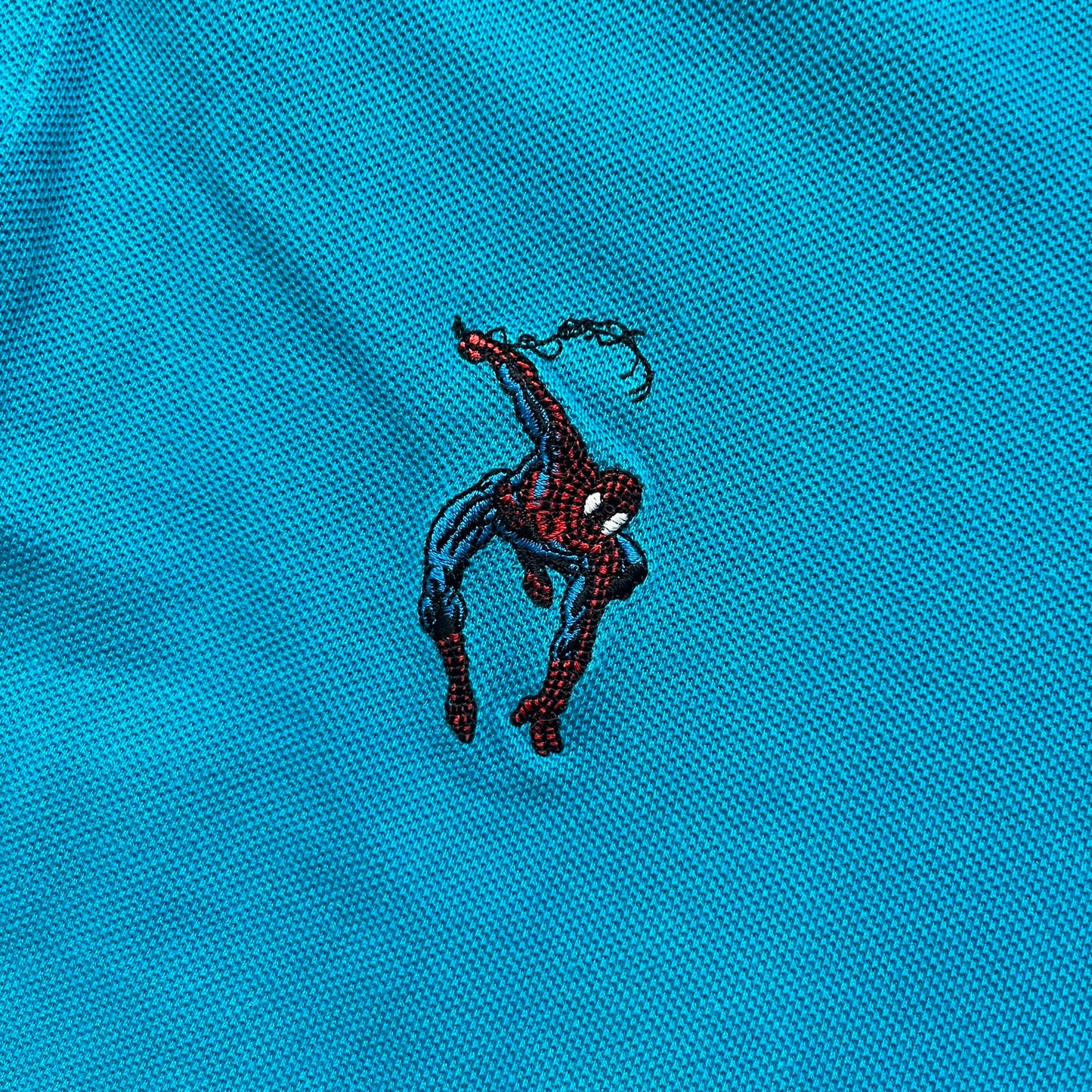 Marvel Comics Spider-Man 1994 Blue Embroidered Polo is a blue polo shirt featuring embroidered Spider-Man branding. The left chest displays an embroidered Spider-Man graphic, while the sleeve includes yellow “Spider-Man” text embroidery. The lower front is finished with small black “1994” text, and the shirt includes a large Marvel Comics neck tag.