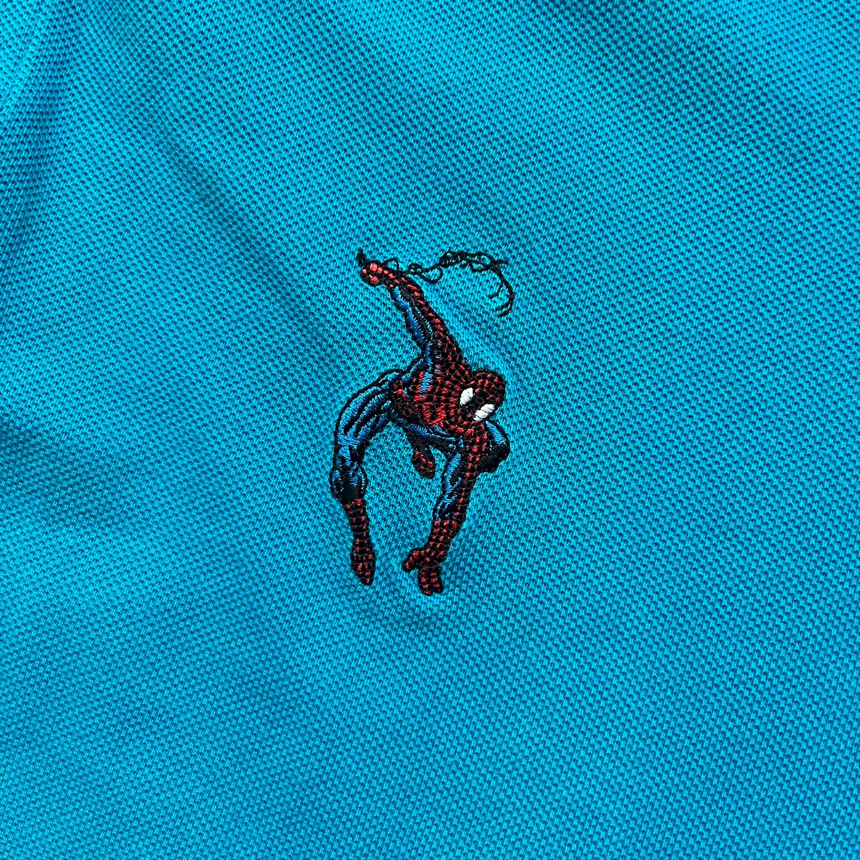 Marvel Comics Spider-Man 1994 Blue Embroidered Polo is a blue polo shirt featuring embroidered Spider-Man branding. The left chest displays an embroidered Spider-Man graphic, while the sleeve includes yellow “Spider-Man” text embroidery. The lower front is finished with small black “1994” text, and the shirt includes a large Marvel Comics neck tag.