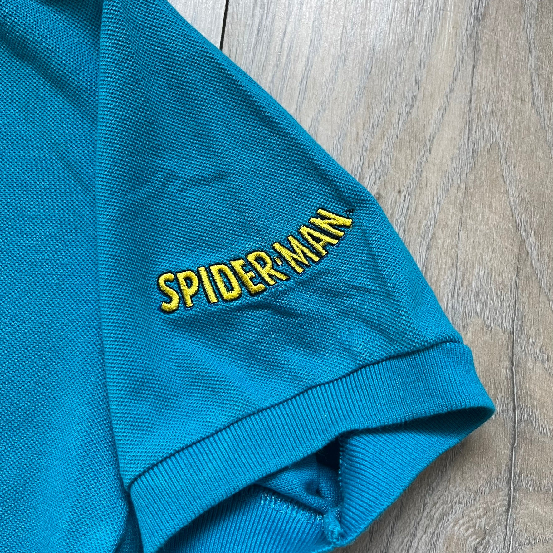 Marvel Comics Spider-Man 1994 Blue Embroidered Polo is a blue polo shirt featuring embroidered Spider-Man branding. The left chest displays an embroidered Spider-Man graphic, while the sleeve includes yellow “Spider-Man” text embroidery. The lower front is finished with small black “1994” text, and the shirt includes a large Marvel Comics neck tag.