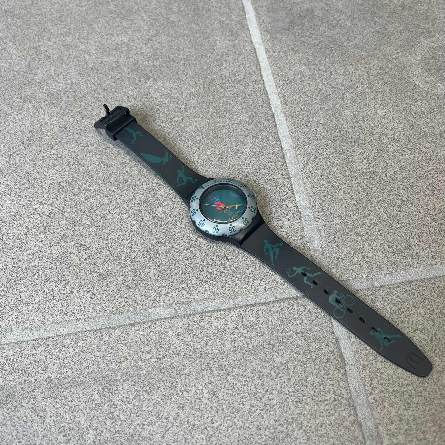 McDonald's 1996 Atlanta Olympics Analog Watch features a black strap decorated with green sports-themed figures and a silver and grey case. The green dial has the Atlanta 1996 Olympic Games logo at the center. A vintage McDonald's promotional watch from the 1996 Atlanta Olympics.
