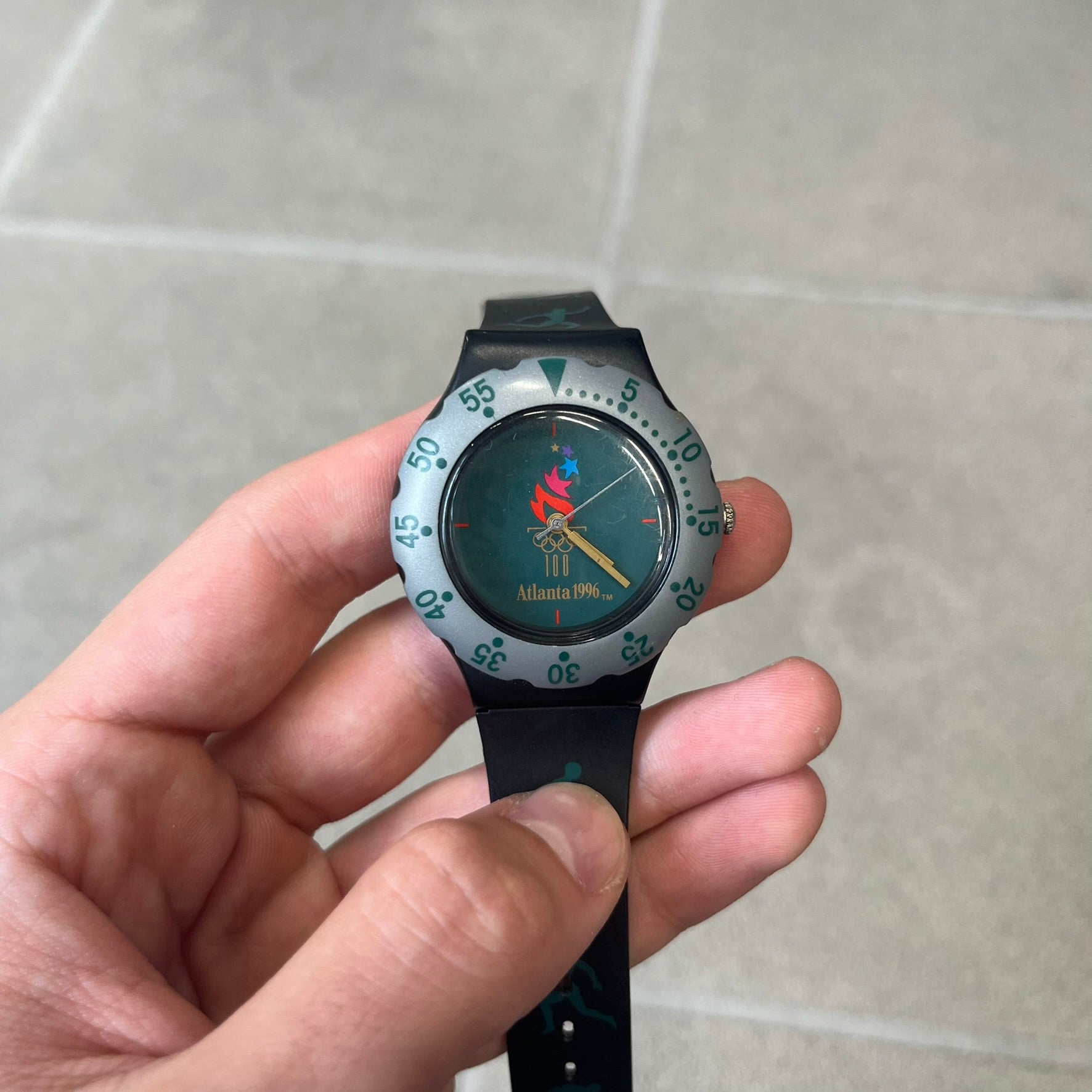 McDonald's 1996 Atlanta Olympics Analog Watch features a black strap decorated with green sports-themed figures and a silver and grey case. The green dial has the Atlanta 1996 Olympic Games logo at the center. A vintage McDonald's promotional watch from the 1996 Atlanta Olympics.
