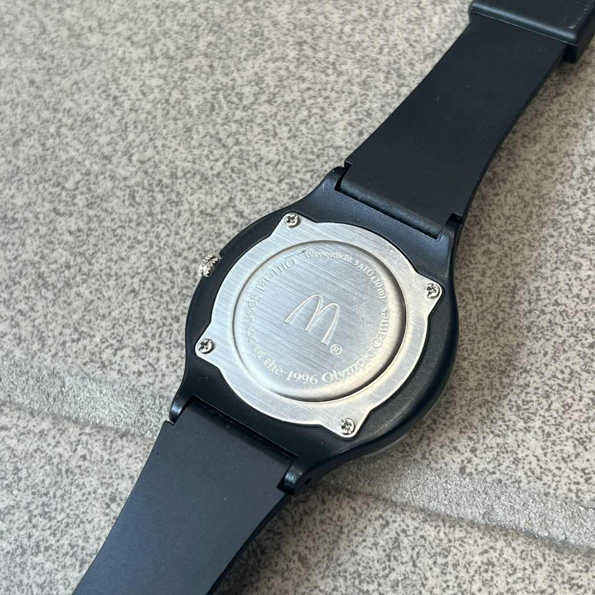 McDonald's 1996 Atlanta Olympics Analog Watch features a black strap decorated with green sports-themed figures and a silver and grey case. The green dial has the Atlanta 1996 Olympic Games logo at the center. A vintage McDonald's promotional watch from the 1996 Atlanta Olympics.
