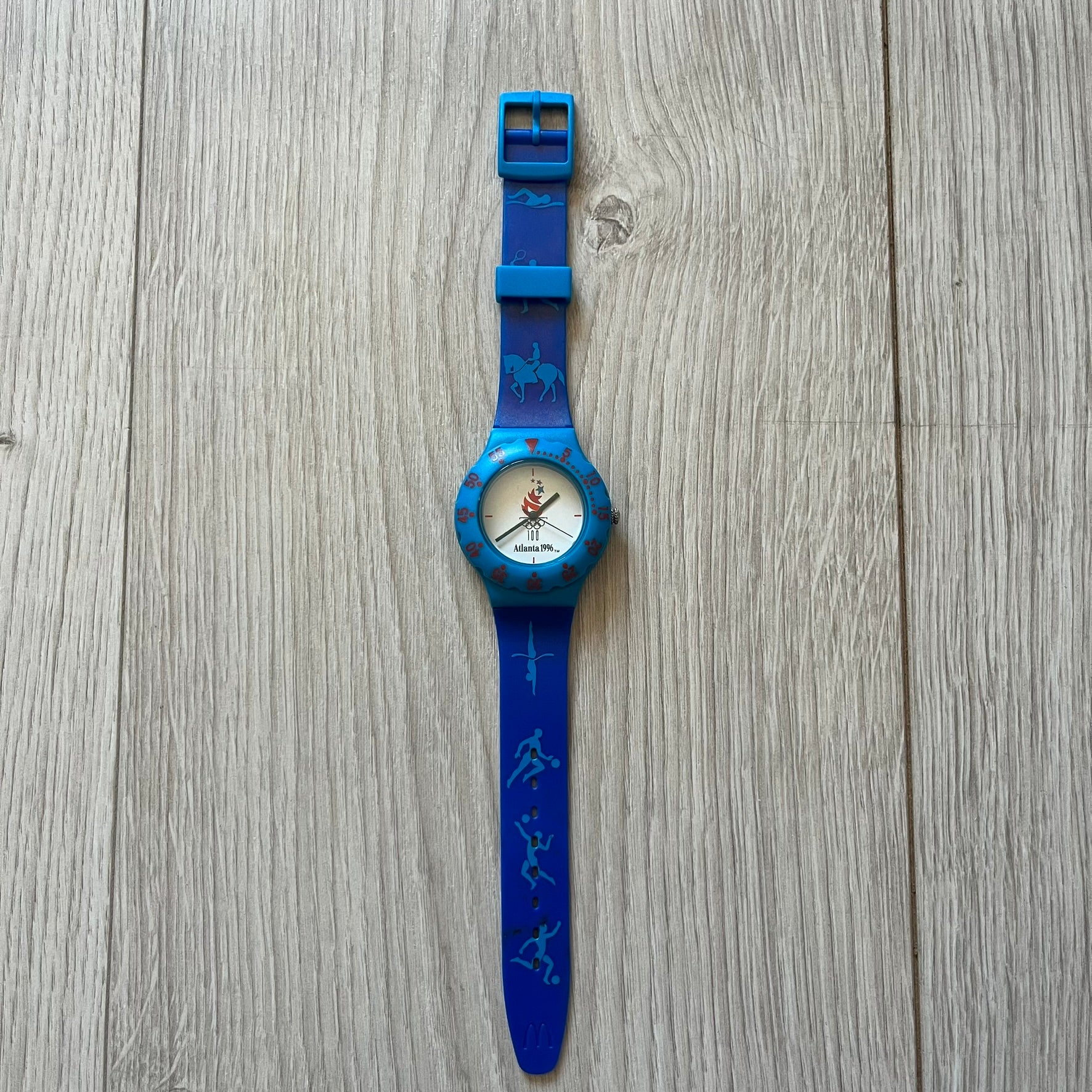 McDonald’s Atlanta 1996 Olympics analog watch with a dark blue strap featuring light blue sport-themed motifs. The dial is white with blue and red accents, showcasing the official Atlanta 1996 logo. A nostalgic collectible celebrating the iconic Olympic collaboration.