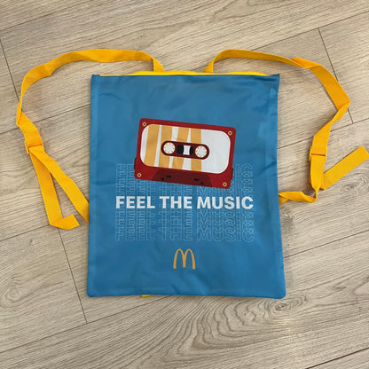 The McDonald’s Spain “Feel The Music” Backpack comes in a blue and yellow colorway with a vintage cassette graphic on the front. Constructed from lightweight non-woven synthetic fabric, it includes yellow straps and a zip-top closure. A collectible promotional item released by McDonald’s Spain.