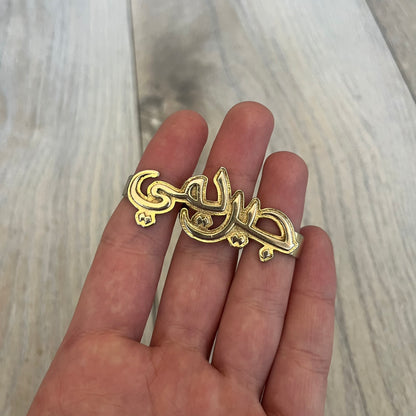 Melody Ehsani x Jeremy Scott 2013 Arabic Three Finger Ring is a gold-tone three-finger ring from the 2013 Fearless collection by Melody Ehsani and Jeremy Scott. The ring features the name “Jeremy” written in Arabic script across the three connected bands. Released in 2013, the piece originally retailed for $100.