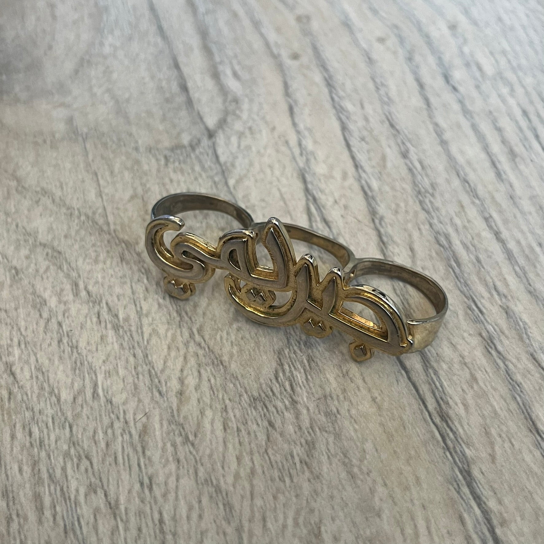 Melody Ehsani x Jeremy Scott 2013 Arabic Three Finger Ring is a gold-tone three-finger ring from the 2013 Fearless collection by Melody Ehsani and Jeremy Scott. The ring features the name “Jeremy” written in Arabic script across the three connected bands. Released in 2013, the piece originally retailed for $100.