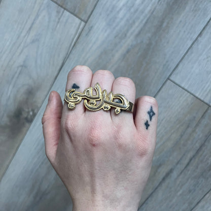 Melody Ehsani x Jeremy Scott 2013 Arabic Three Finger Ring is a gold-tone three-finger ring from the 2013 Fearless collection by Melody Ehsani and Jeremy Scott. The ring features the name “Jeremy” written in Arabic script across the three connected bands. Released in 2013, the piece originally retailed for $100.
