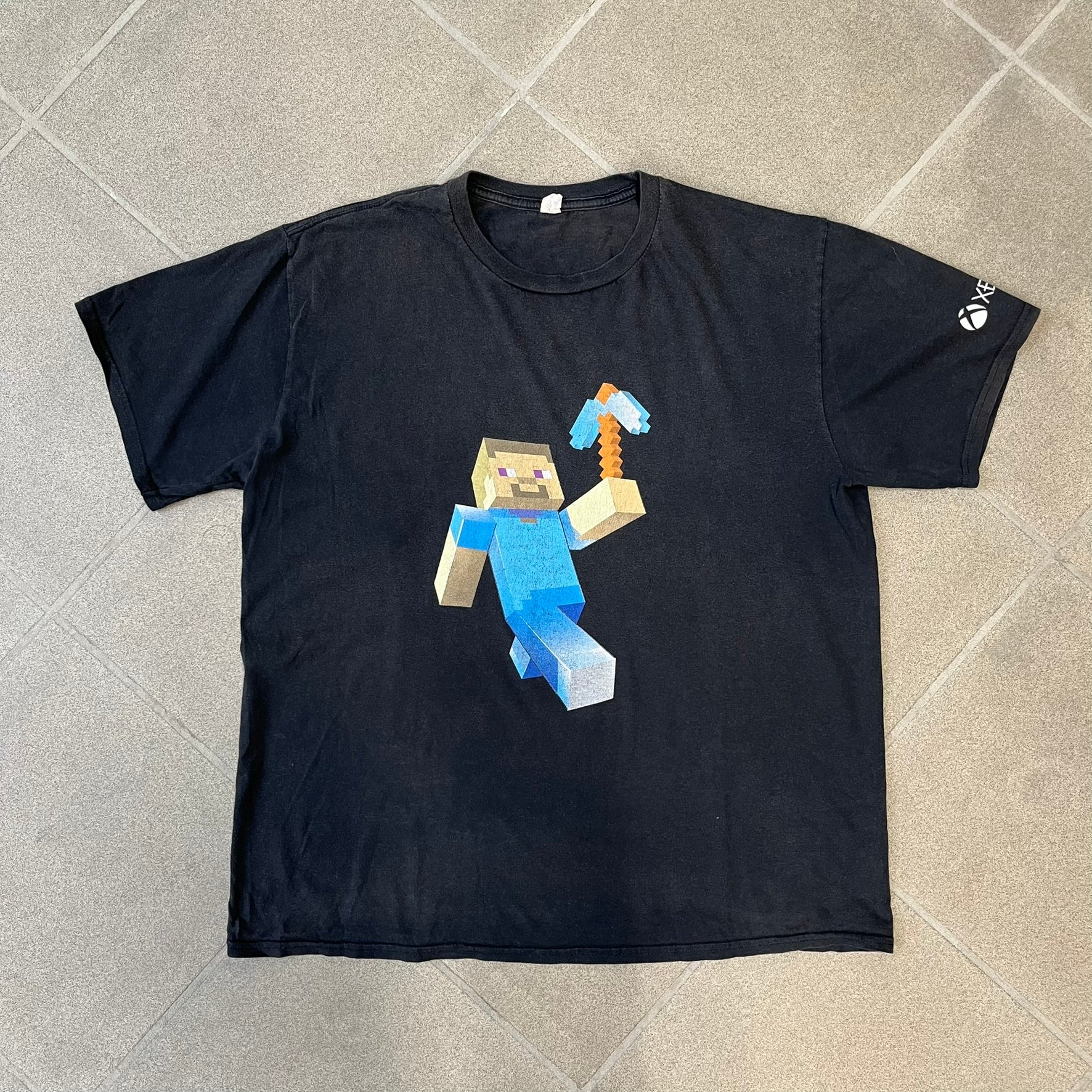 Minecraft 2012 Xbox 360 Black Promo Tee features a black design with Steve from Minecraft walking while holding a diamond pickaxe on the front. The sleeve has a white Xbox 360 logo, while the back features three white gaming-related logos including Mojang. A promotional release from 2012.

