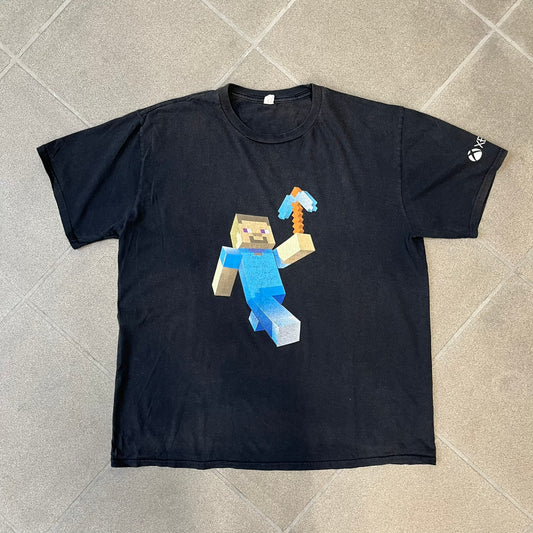 Minecraft 2012 Xbox 360 Black Promo Tee features a black design with Steve from Minecraft walking while holding a diamond pickaxe on the front. The sleeve has a white Xbox 360 logo, while the back features three white gaming-related logos including Mojang. A promotional release from 2012.
