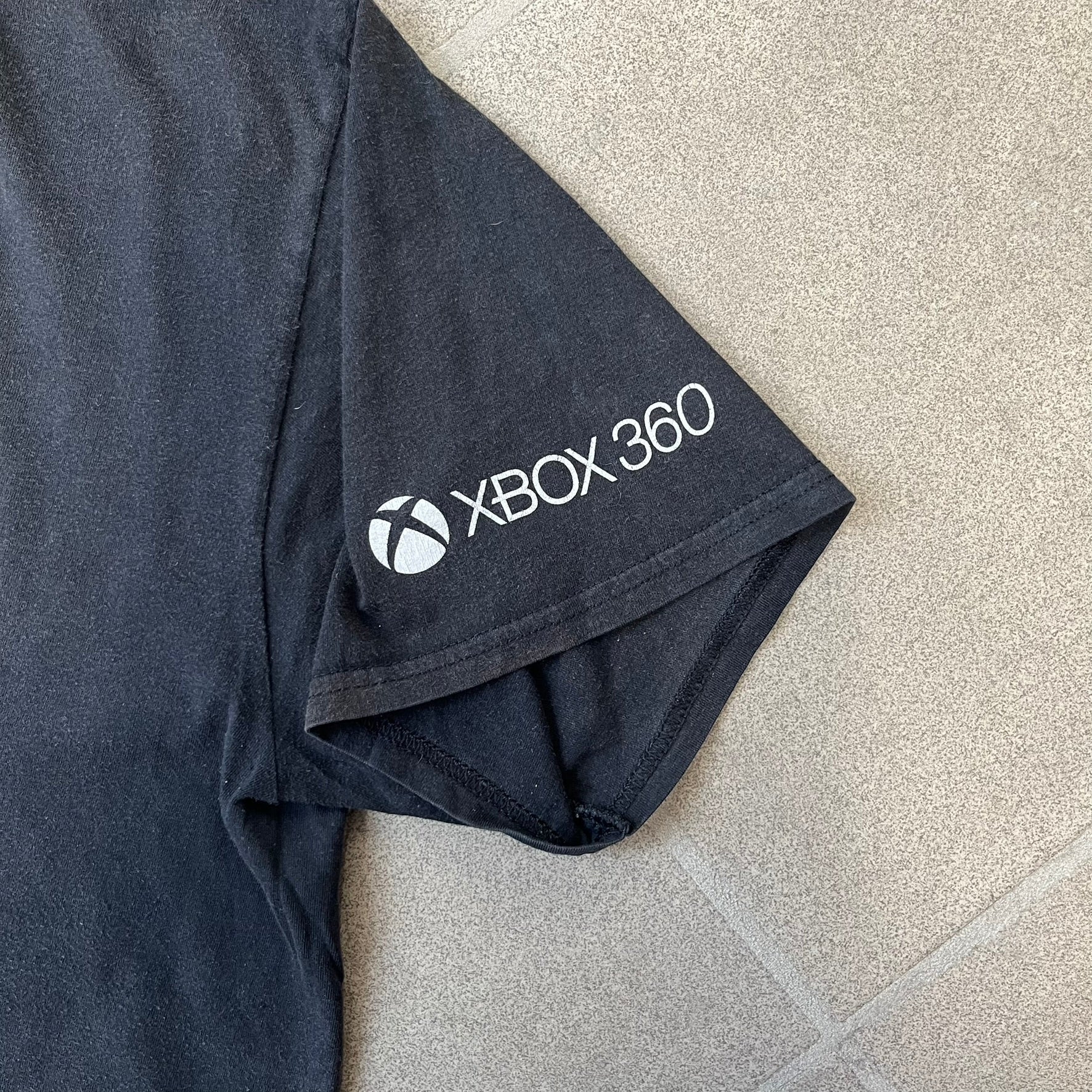 Minecraft 2012 Xbox 360 Black Promo Tee features a black design with Steve from Minecraft walking while holding a diamond pickaxe on the front. The sleeve has a white Xbox 360 logo, while the back features three white gaming-related logos including Mojang. A promotional release from 2012.
