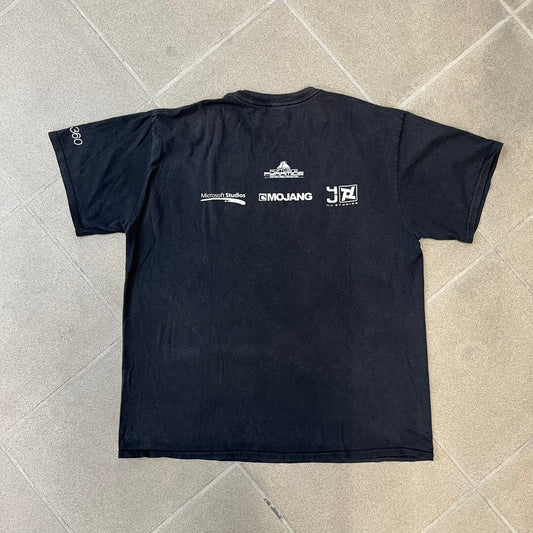 Minecraft 2012 Xbox 360 Black Promo Tee features a black design with Steve from Minecraft walking while holding a diamond pickaxe on the front. The sleeve has a white Xbox 360 logo, while the back features three white gaming-related logos including Mojang. A promotional release from 2012.

