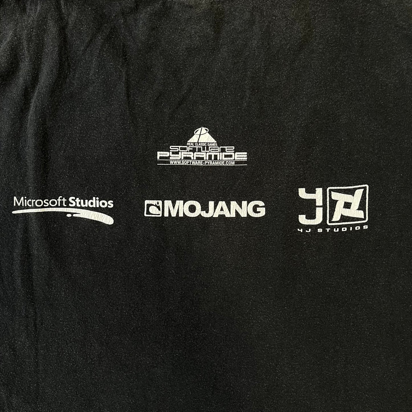 Minecraft 2012 Xbox 360 Black Promo Tee features a black design with Steve from Minecraft walking while holding a diamond pickaxe on the front. The sleeve has a white Xbox 360 logo, while the back features three white gaming-related logos including Mojang. A promotional release from 2012.
