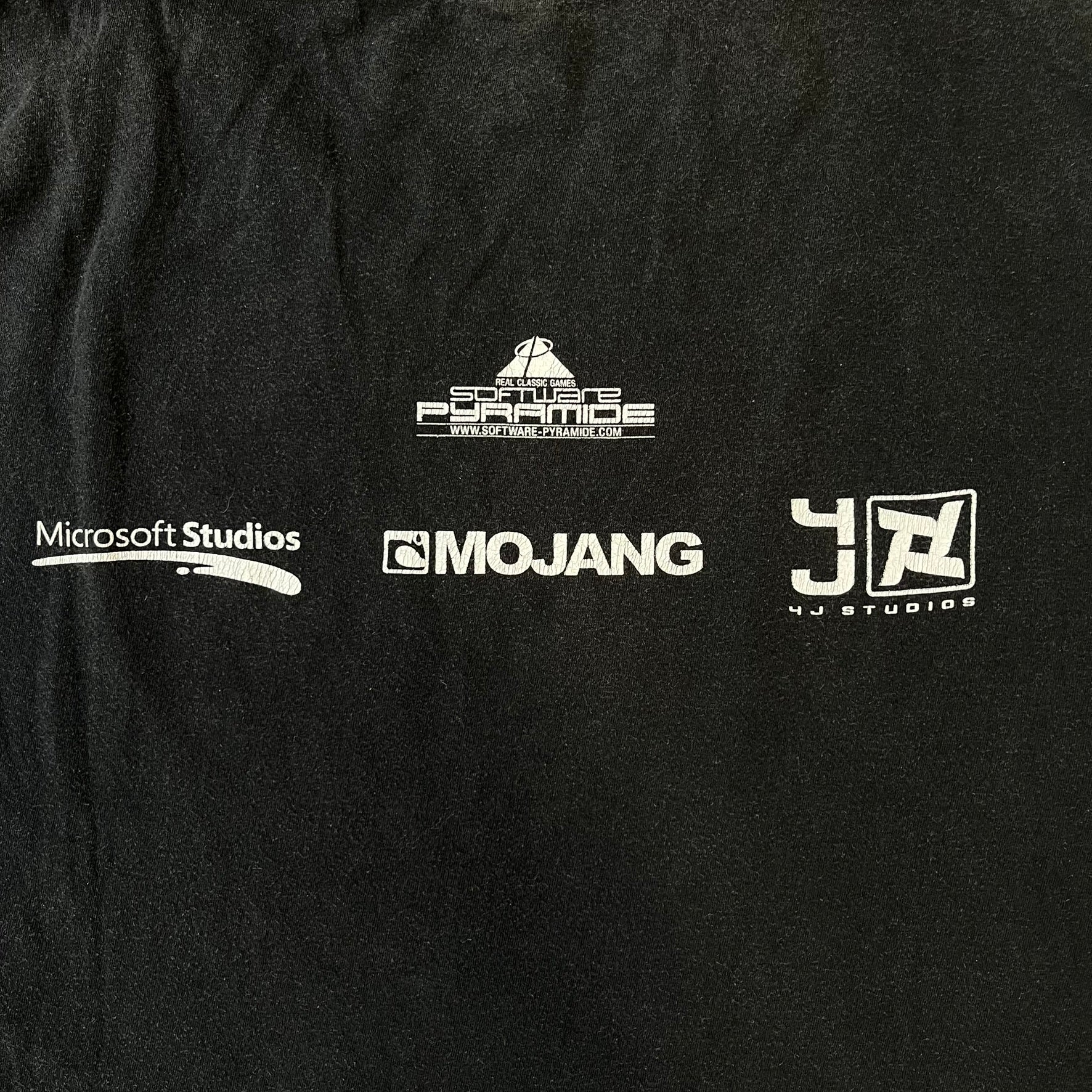 Minecraft 2012 Xbox 360 Black Promo Tee features a black design with Steve from Minecraft walking while holding a diamond pickaxe on the front. The sleeve has a white Xbox 360 logo, while the back features three white gaming-related logos including Mojang. A promotional release from 2012.

