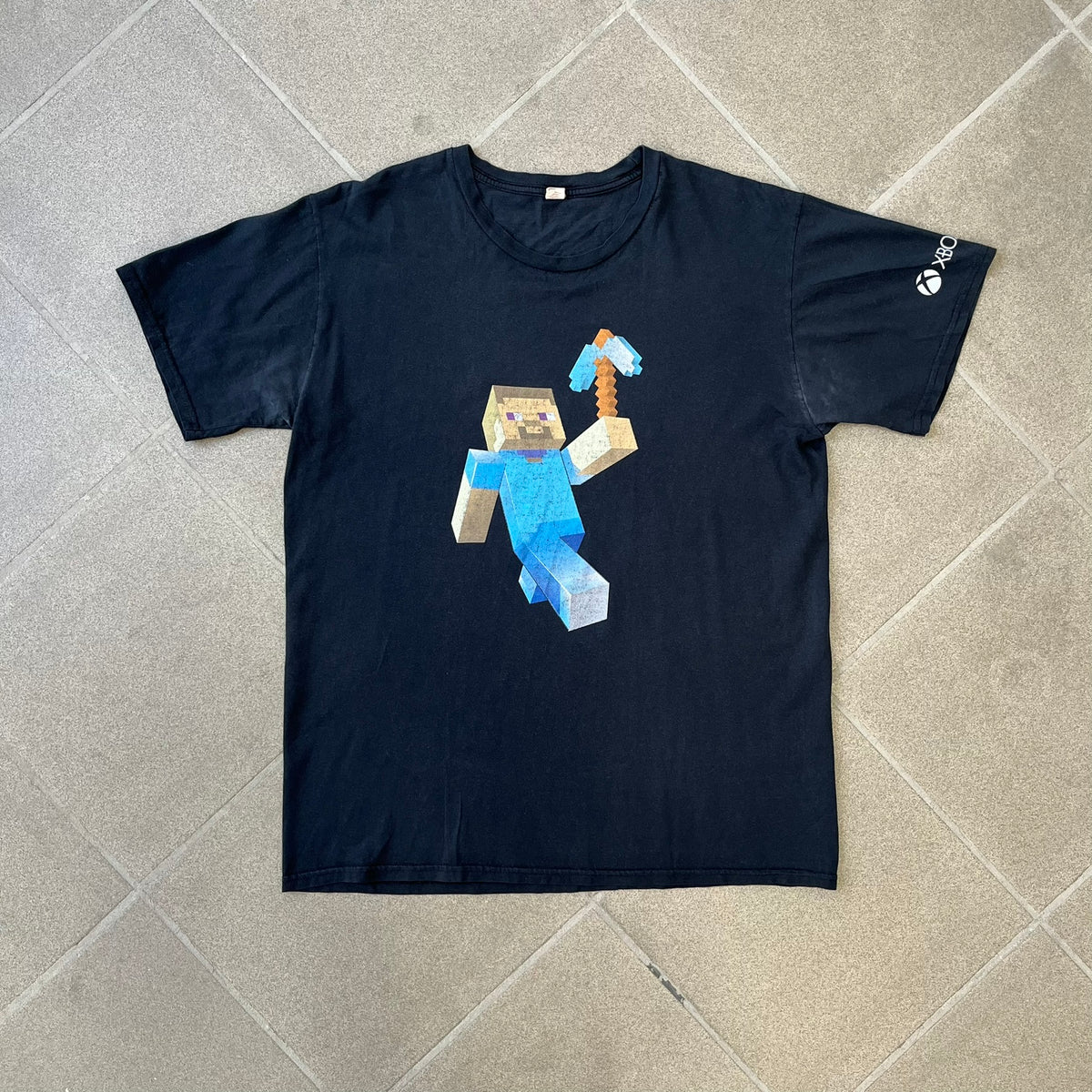 Minecraft 2012 Xbox 360 Black Promo Tee features a black design with Steve from Minecraft walking while holding a diamond pickaxe on the front. The sleeve has a white Xbox 360 logo, while the back features three white gaming-related logos including Mojang. A promotional release from 2012.
