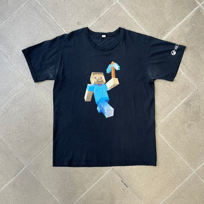 Minecraft 2012 Xbox 360 Black Promo Tee features a black design with Steve from Minecraft walking while holding a diamond pickaxe on the front. The sleeve has a white Xbox 360 logo, while the back features three white gaming-related logos including Mojang. A promotional release from 2012.
