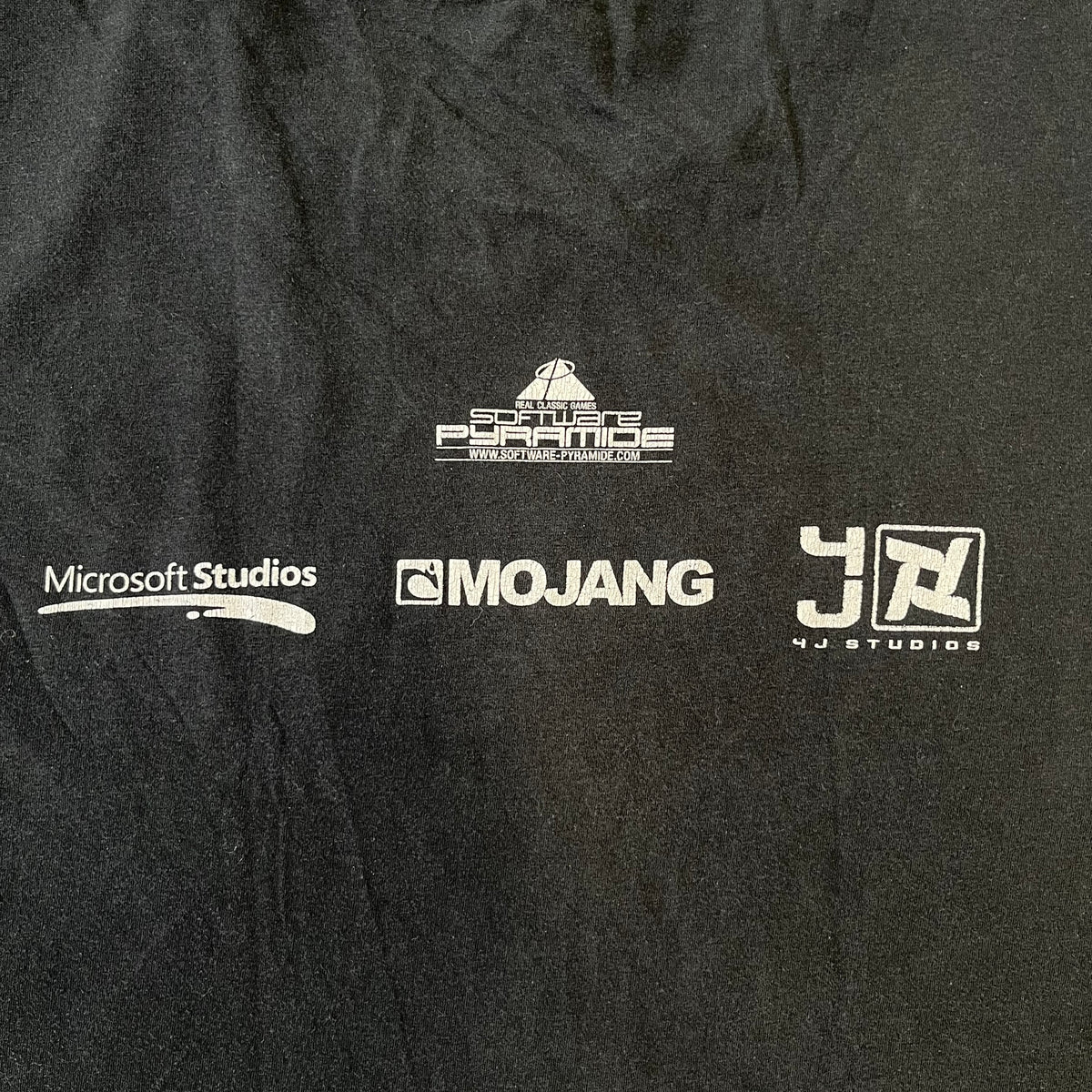 Minecraft 2012 Xbox 360 Black Promo Tee features a black design with Steve from Minecraft walking while holding a diamond pickaxe on the front. The sleeve has a white Xbox 360 logo, while the back features three white gaming-related logos including Mojang. A promotional release from 2012.
