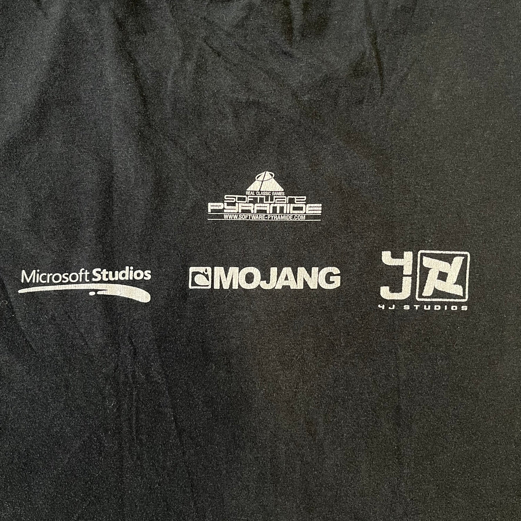 Minecraft 2012 Xbox 360 Black Promo Tee features a black design with Steve from Minecraft walking while holding a diamond pickaxe on the front. The sleeve has a white Xbox 360 logo, while the back features three white gaming-related logos including Mojang. A promotional release from 2012.
