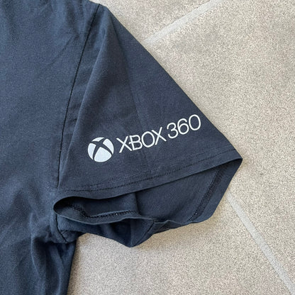 Minecraft 2012 Xbox 360 Black Promo Tee features a black design with Steve from Minecraft walking while holding a diamond pickaxe on the front. The sleeve has a white Xbox 360 logo, while the back features three white gaming-related logos including Mojang. A promotional release from 2012.
