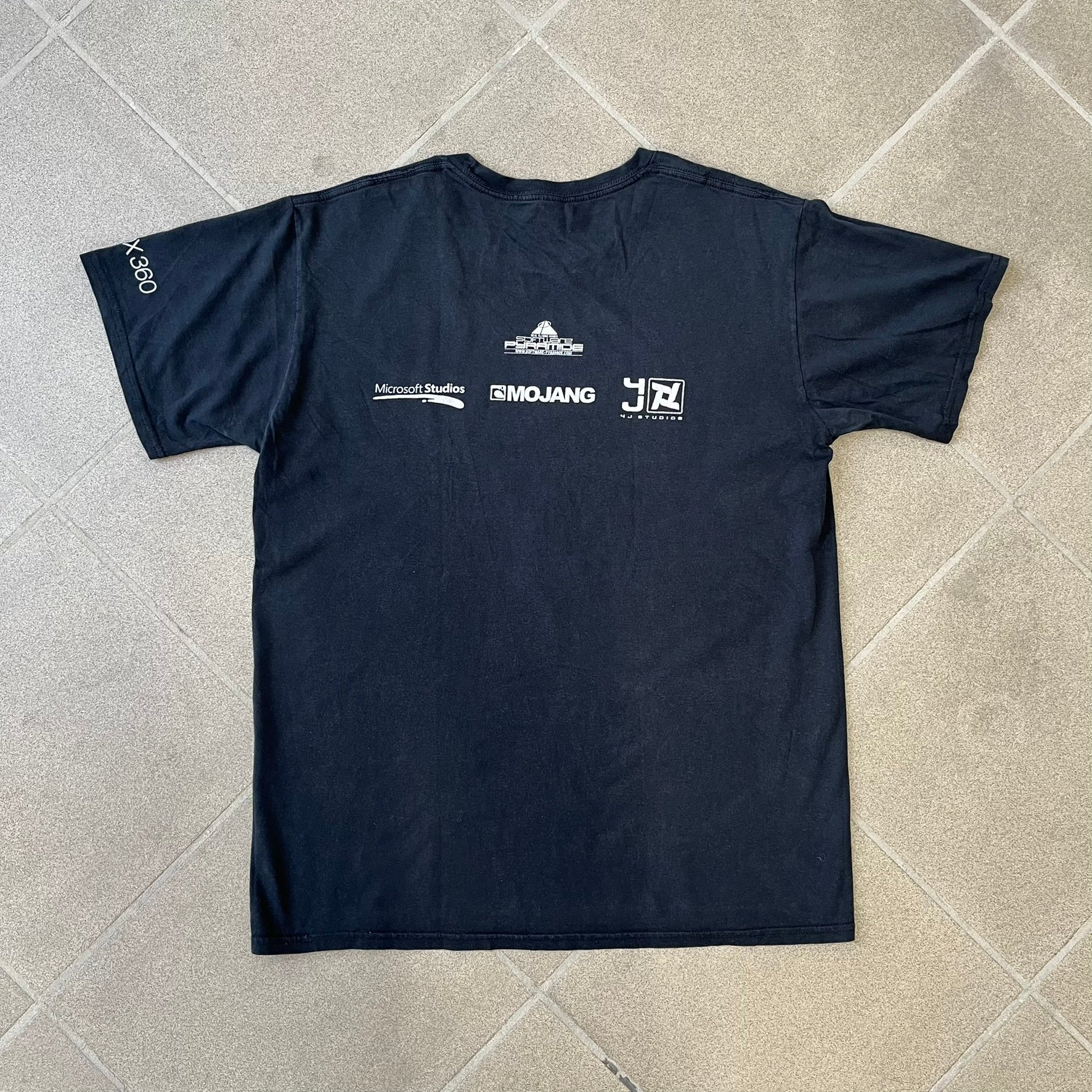 Minecraft 2012 Xbox 360 Black Promo Tee features a black design with Steve from Minecraft walking while holding a diamond pickaxe on the front. The sleeve has a white Xbox 360 logo, while the back features three white gaming-related logos including Mojang. A promotional release from 2012.
