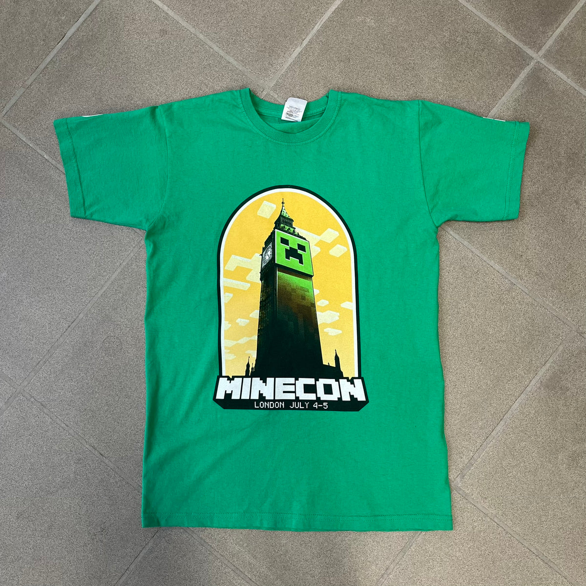 Minecraft 2015 Minecon London Green Tee is a green T-shirt made for Minecon 2015 in London, featuring Big Ben with a Creeper face on the front, with “Minecon London July 4-5” text in Minecraft-style lettering. The upper back displays “Minecon” with “Thanks for Playing” text below, while the sleeves carry a Mojang logo on one side and a Microsoft logo on the other.