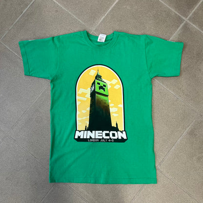 Minecraft 2015 Minecon London Green Tee is a green T-shirt made for Minecon 2015 in London, featuring Big Ben with a Creeper face on the front, with “Minecon London July 4-5” text in Minecraft-style lettering. The upper back displays “Minecon” with “Thanks for Playing” text below, while the sleeves carry a Mojang logo on one side and a Microsoft logo on the other.