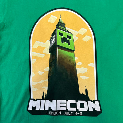 Minecraft 2015 Minecon London Green Tee is a green T-shirt made for Minecon 2015 in London, featuring Big Ben with a Creeper face on the front, with “Minecon London July 4-5” text in Minecraft-style lettering. The upper back displays “Minecon” with “Thanks for Playing” text below, while the sleeves carry a Mojang logo on one side and a Microsoft logo on the other.