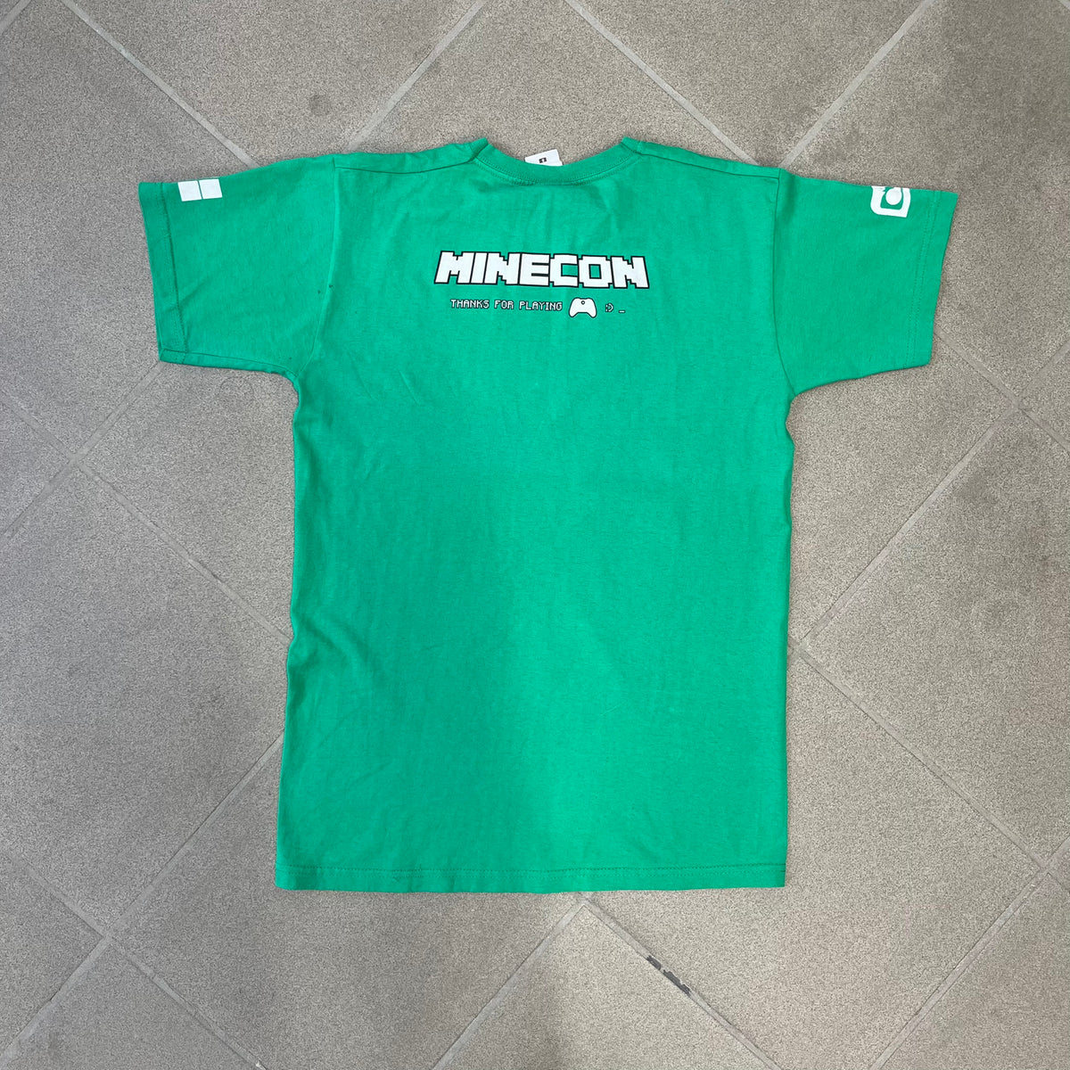 Minecraft 2015 Minecon London Green Tee is a green T-shirt made for Minecon 2015 in London, featuring Big Ben with a Creeper face on the front, with “Minecon London July 4-5” text in Minecraft-style lettering. The upper back displays “Minecon” with “Thanks for Playing” text below, while the sleeves carry a Mojang logo on one side and a Microsoft logo on the other.