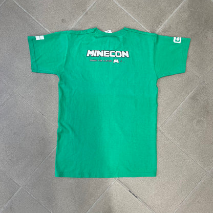 Minecraft 2015 Minecon London Green Tee is a green T-shirt made for Minecon 2015 in London, featuring Big Ben with a Creeper face on the front, with “Minecon London July 4-5” text in Minecraft-style lettering. The upper back displays “Minecon” with “Thanks for Playing” text below, while the sleeves carry a Mojang logo on one side and a Microsoft logo on the other.