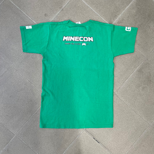Minecraft 2015 Minecon London Green Tee is a green T-shirt made for Minecon 2015 in London, featuring Big Ben with a Creeper face on the front, with “Minecon London July 4-5” text in Minecraft-style lettering. The upper back displays “Minecon” with “Thanks for Playing” text below, while the sleeves carry a Mojang logo on one side and a Microsoft logo on the other.