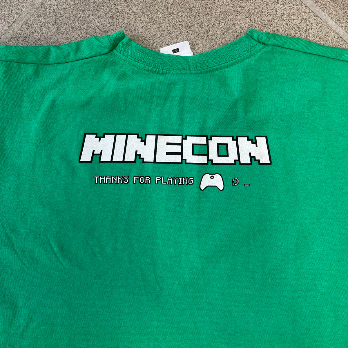 Minecraft 2015 Minecon London Green Tee is a green T-shirt made for Minecon 2015 in London, featuring Big Ben with a Creeper face on the front, with “Minecon London July 4-5” text in Minecraft-style lettering. The upper back displays “Minecon” with “Thanks for Playing” text below, while the sleeves carry a Mojang logo on one side and a Microsoft logo on the other.